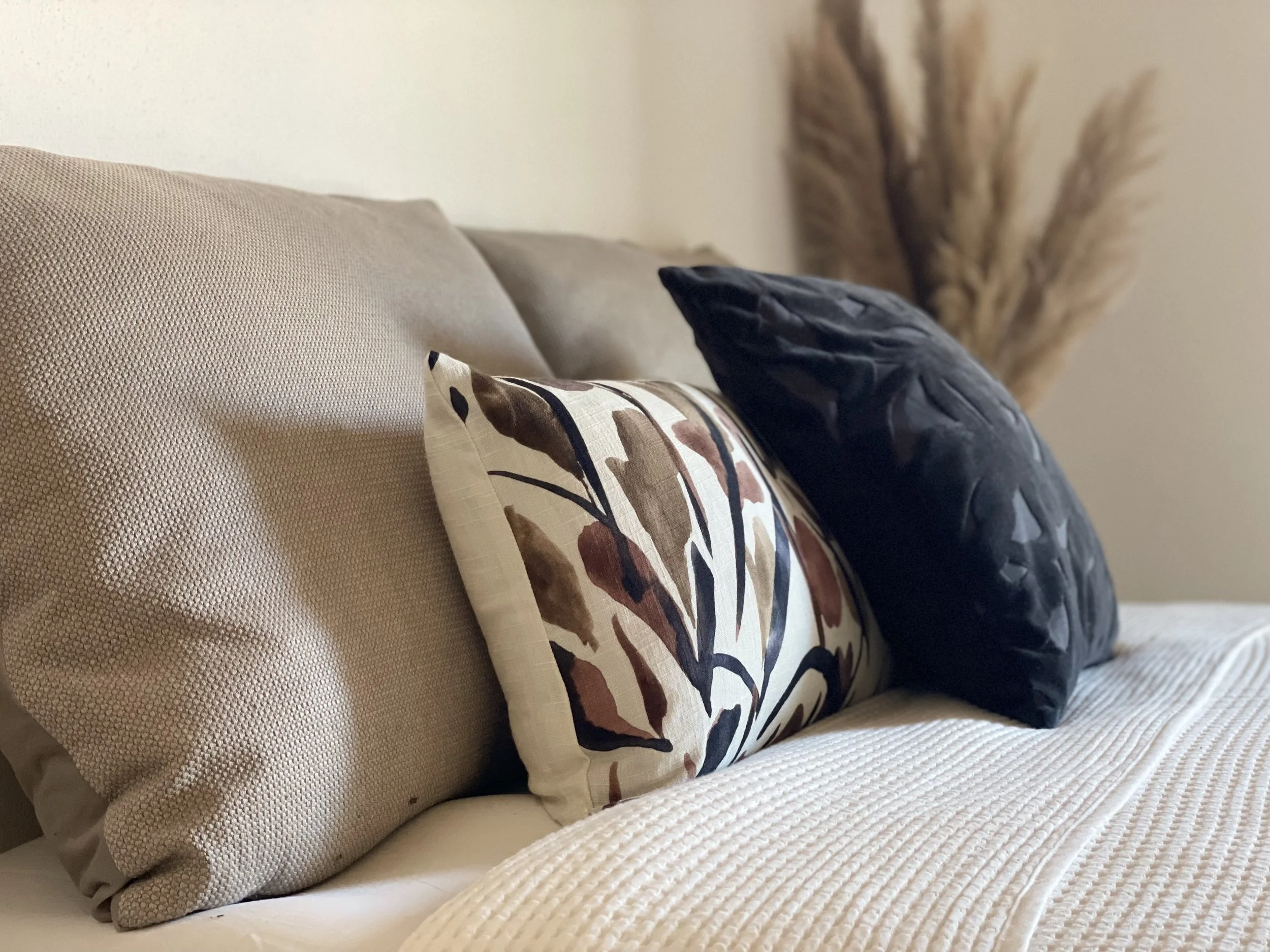 Cuscini decorativi su un letto beige con coperte bianche, sfondo di piante secche.