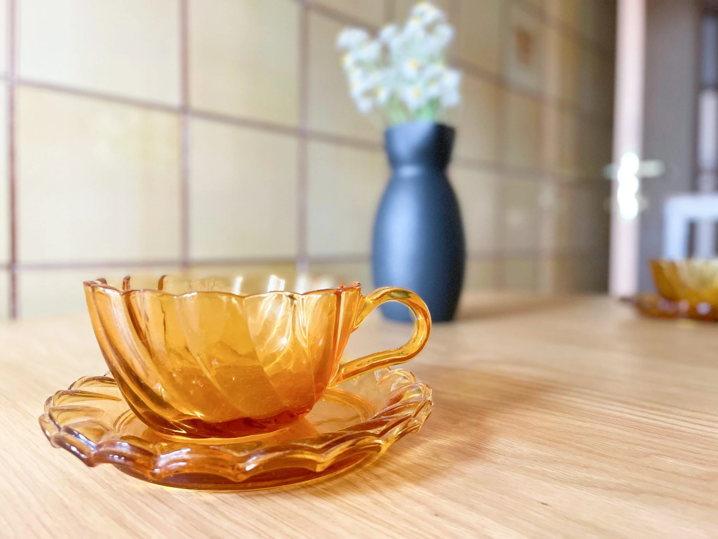 Tazza di vetro ambrato con piattino su un tavolo di legno, sfondo con vaso nero e fiori bianchi.