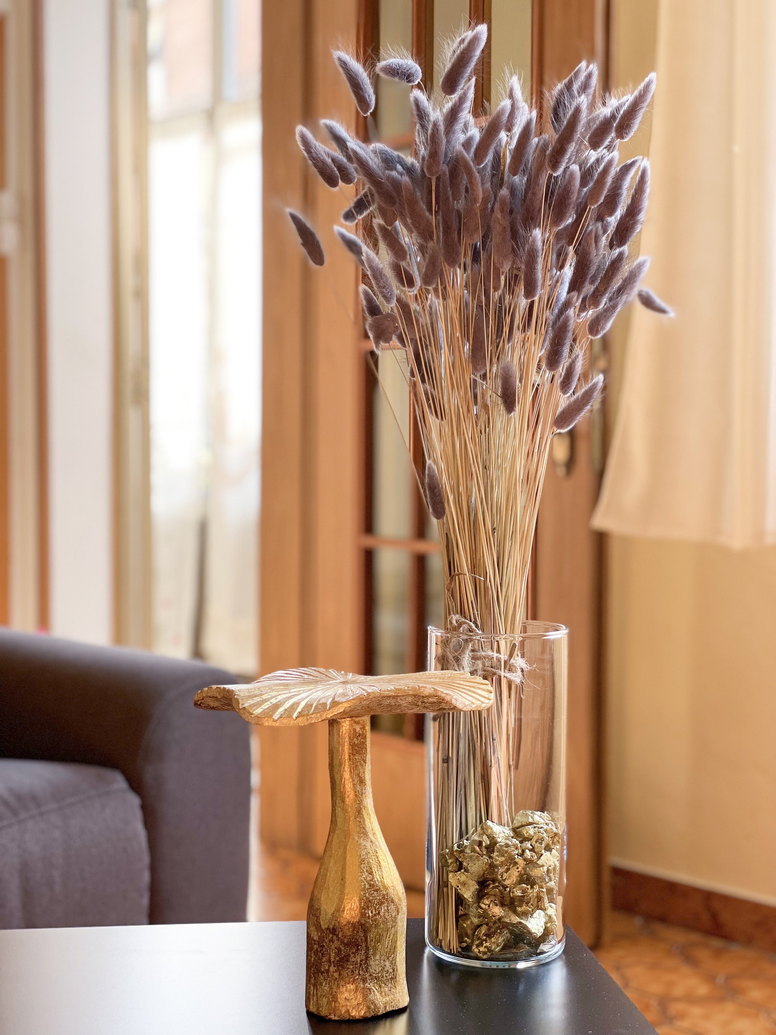 Un vaso trasparente contenente fiori secchi di lagurus su un tavolo. Accanto, una scultura decorativa in legno dorato. Sullo sfondo, un divano e una finestra con tenda in una stanza luminosa.