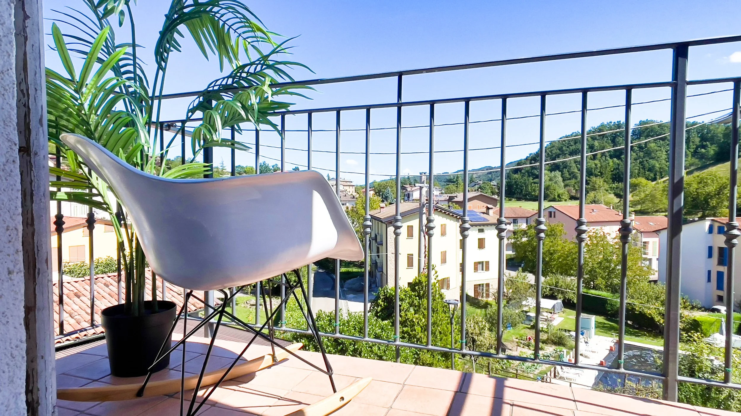 Una sedia bianca su un balcone con una pianta in vaso e vista su edifici e colline verdi.