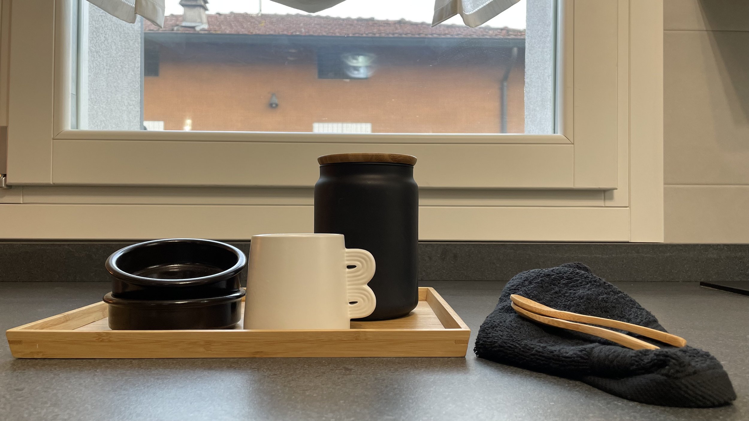 Un davanzale di cucina con un vassoio di legno contenente tazze nere e bianche, un barattolo nero con coperchio di legno, e un asciugamano nero con un cucchiaio di legno accanto, con vista su una finestra.