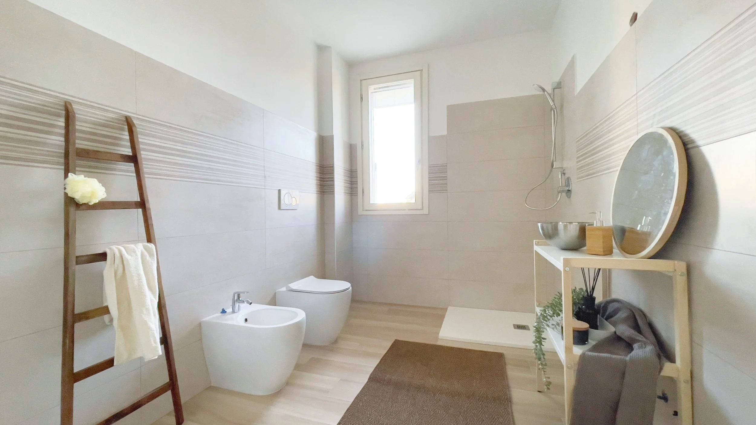 Bagno moderno con piastrelle beige, una scala in legno per asciugamani, bidet, wc, cabina doccia, specchio rotondo su tavolo con lavabo.