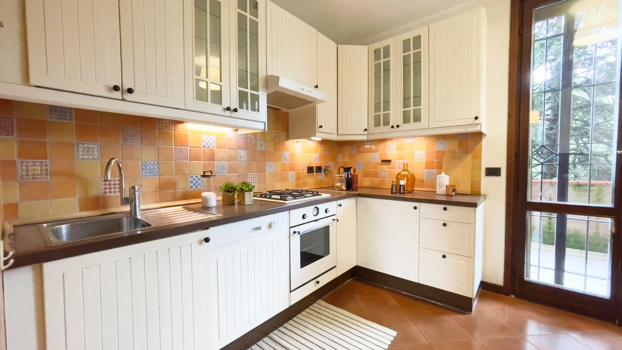 Cucina moderna con mobili bianchi, piano cottura a gas, lavandino, piastrelle colorate sullo sfondo, e una finestra con vista sul giardino.