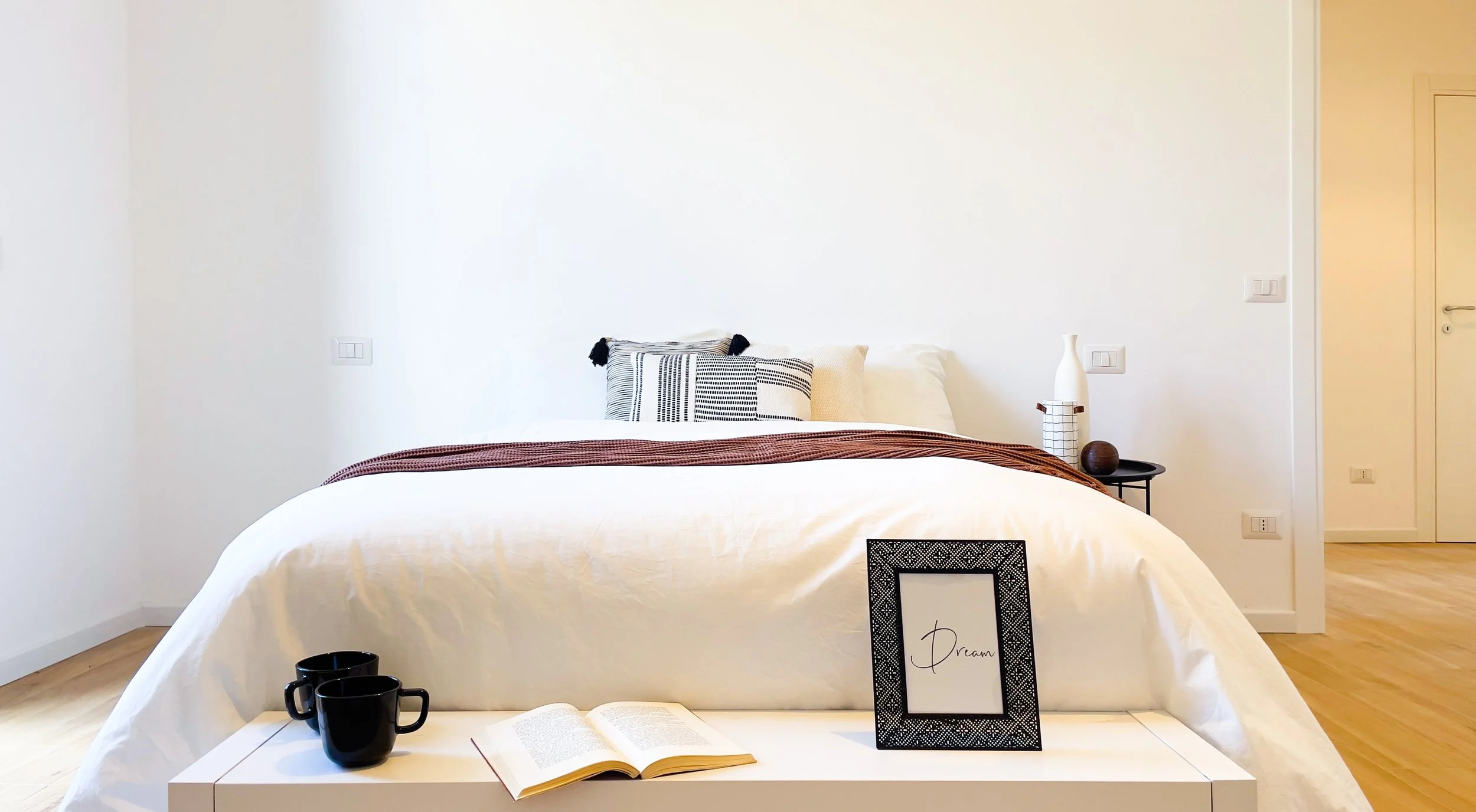 Camera da letto minimalista con letto matrimoniale, cuscini decorativi, coperta marrone, tavolino con decorazioni e libro aperto su un comodino.