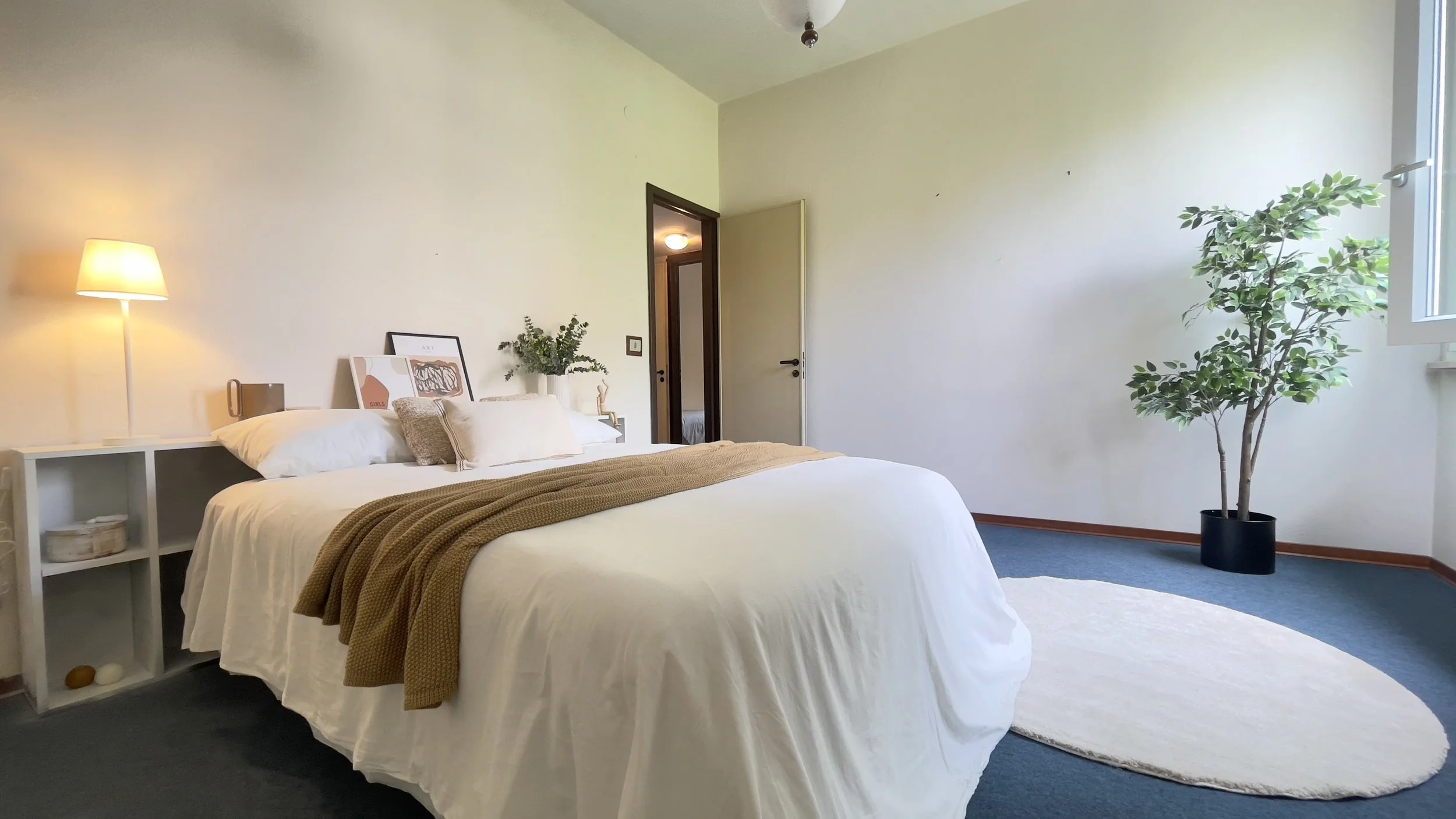 Camera da letto moderna con letto bianco, coperta marrone, lampada da tavolo, piante, tappeto rotondo chiaro e finestra luminosa.