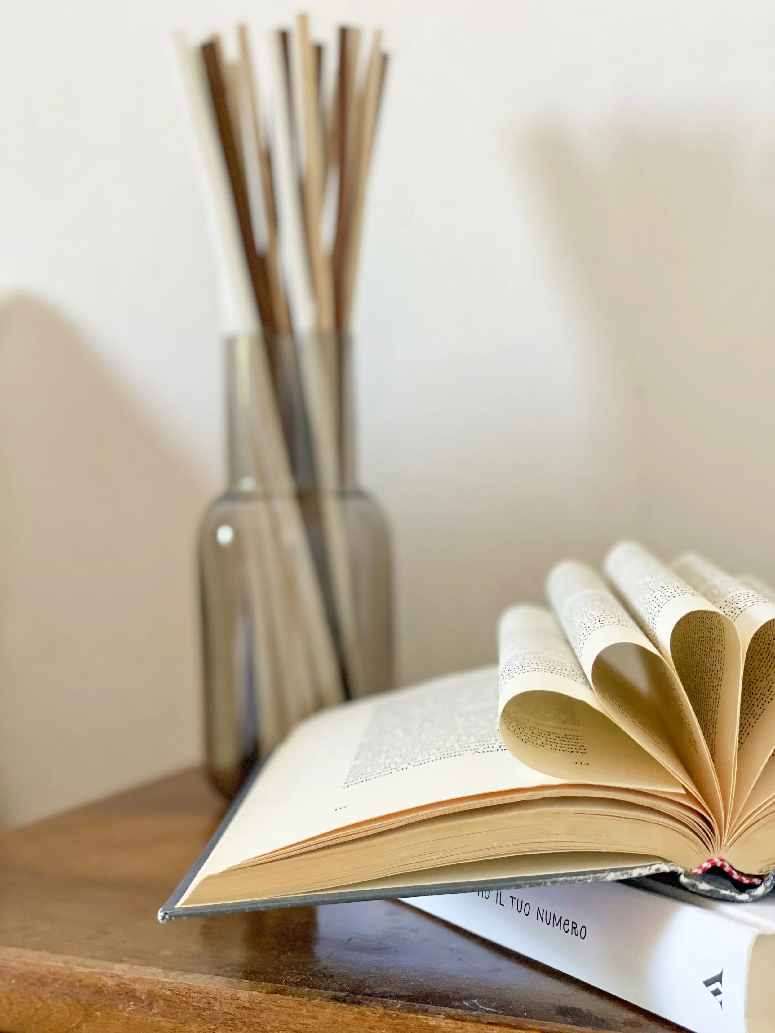Libro aperto su un tavolo di legno con alcune pagine piegate a forma di cuore e un vaso di vetro con bastoncini decorativi sullo sfondo.