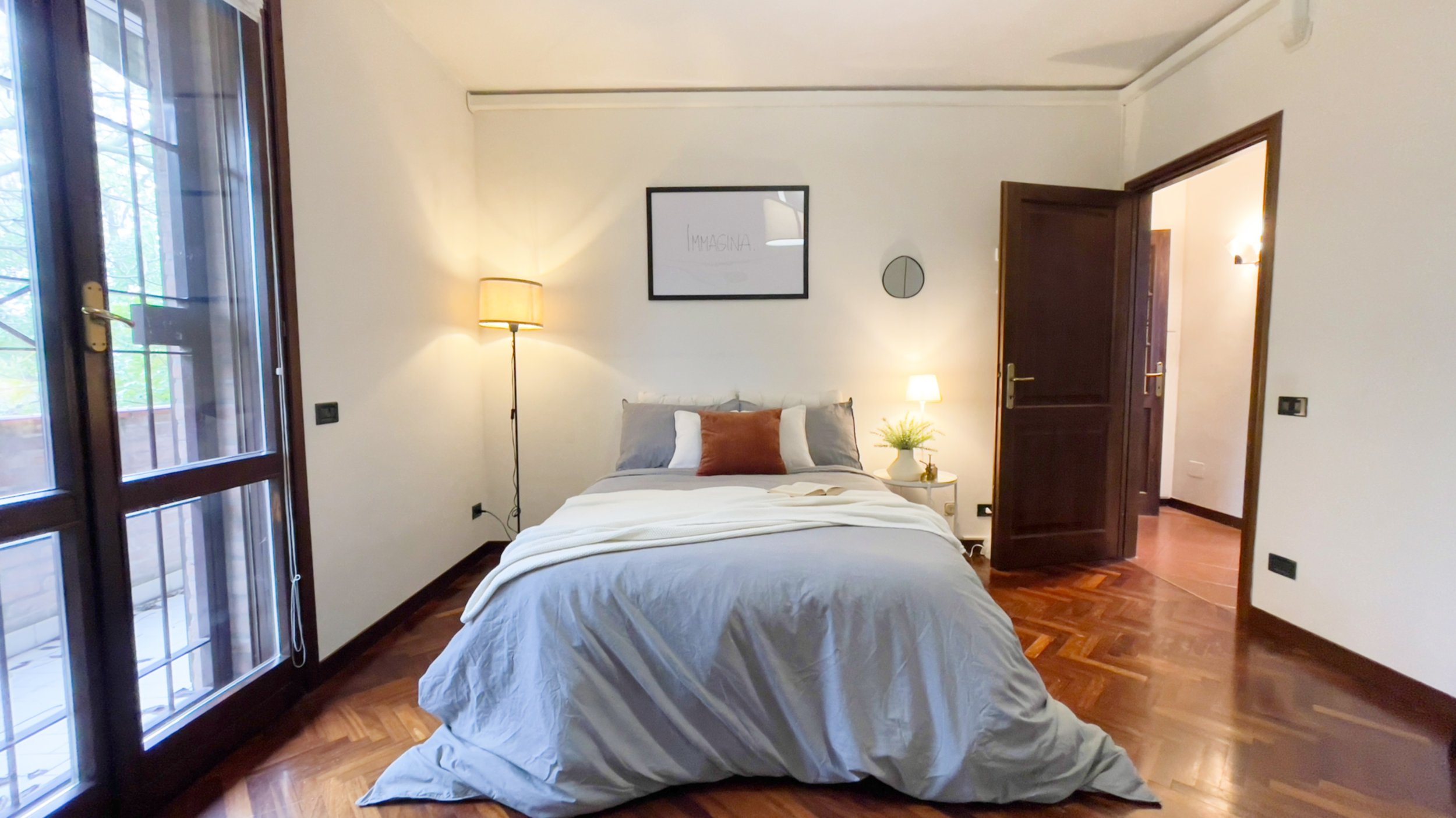 Camera da letto moderna con letto matrimoniale, lenzuola grigie, cuscini decorativi, lampada da terra, comodino con lampada e pianta, porte-finestre che danno su un balcone e pavimento in legno parquet.
