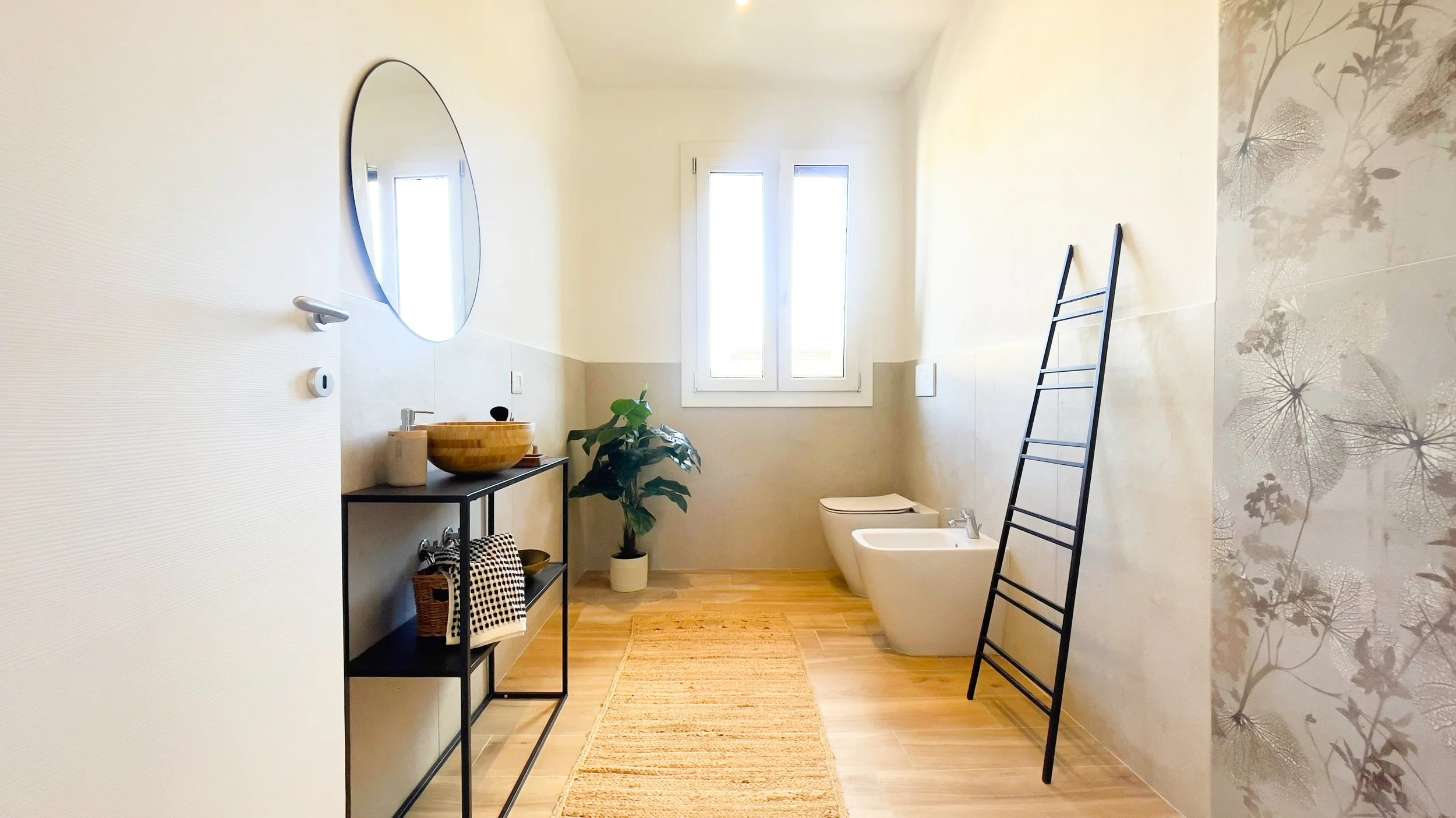 Bagno moderno luminoso con specchio rotondo, lavandino con mobile nero, pianta ornamentale, finestra, bidet e WC, scala nera decorativa e carta da parati floreale.