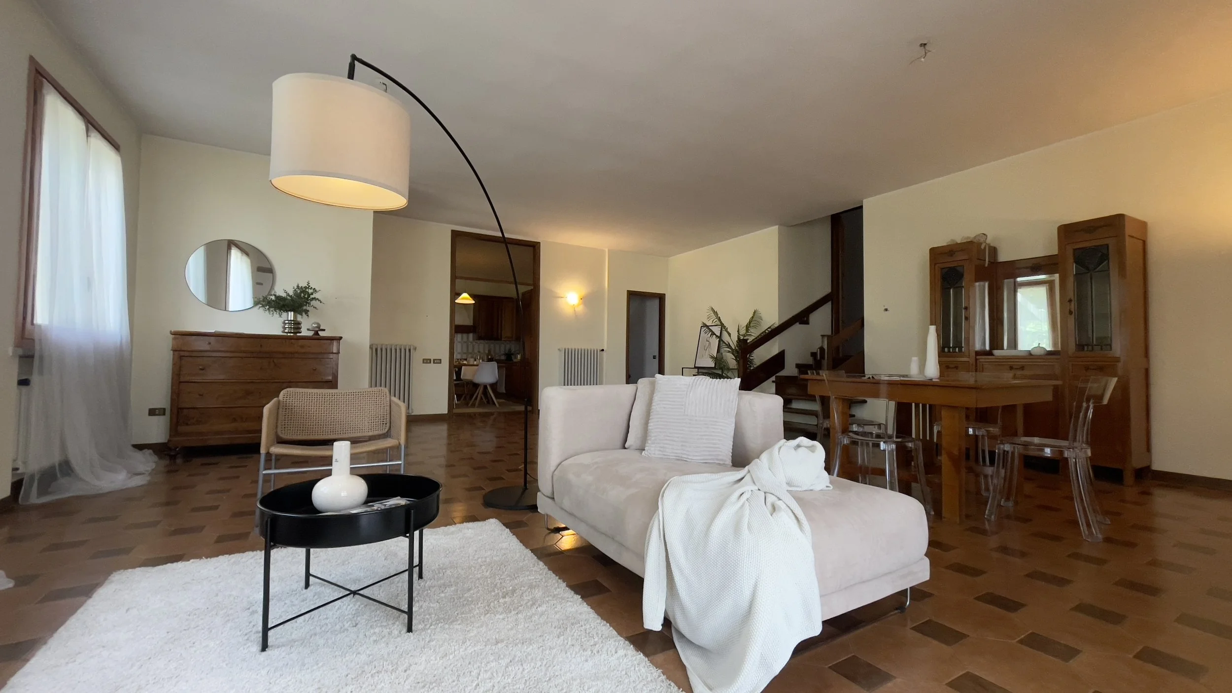 Soggiorno arredato con divano beige, tavolino nero, lampada ad arco, tappeto bianco, mobili in legno, tavolo da pranzo con sedie trasparenti, e finestra con tende bianche.