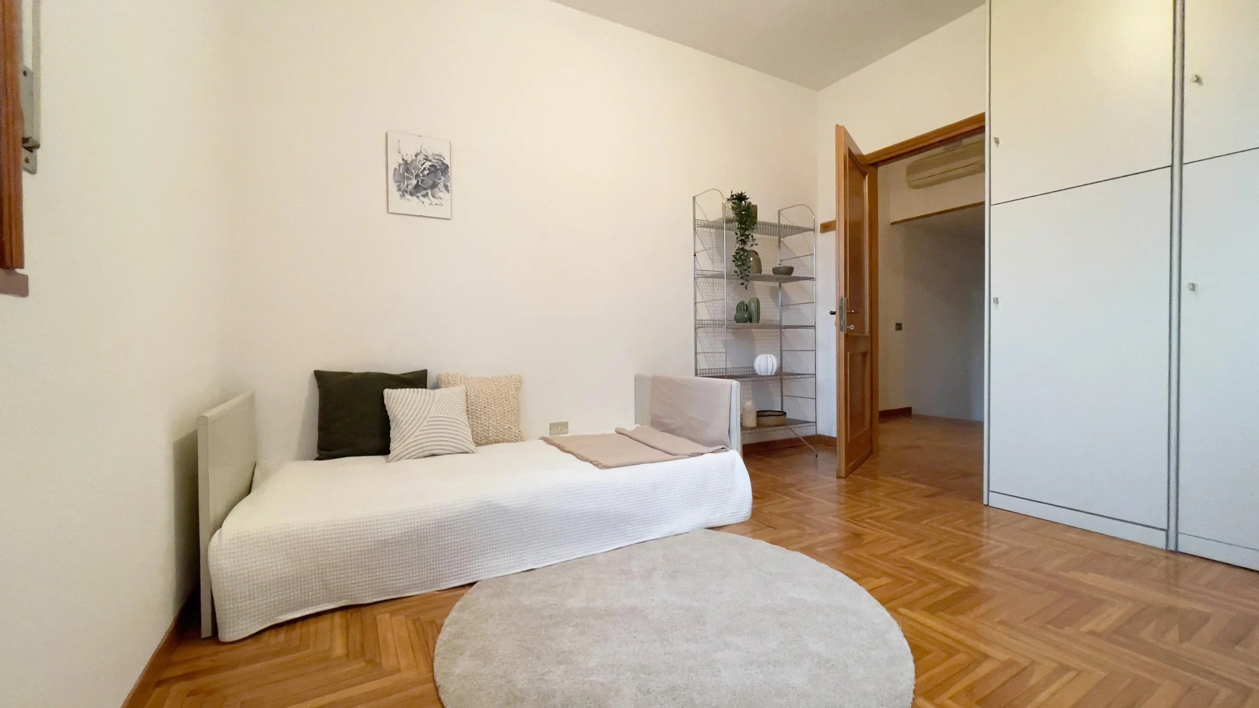 Stanza minimalista con letto singolo e cuscini, scaffale metallico con decorazioni, porta aperta, pavimento in legno e tappeto rotondo.