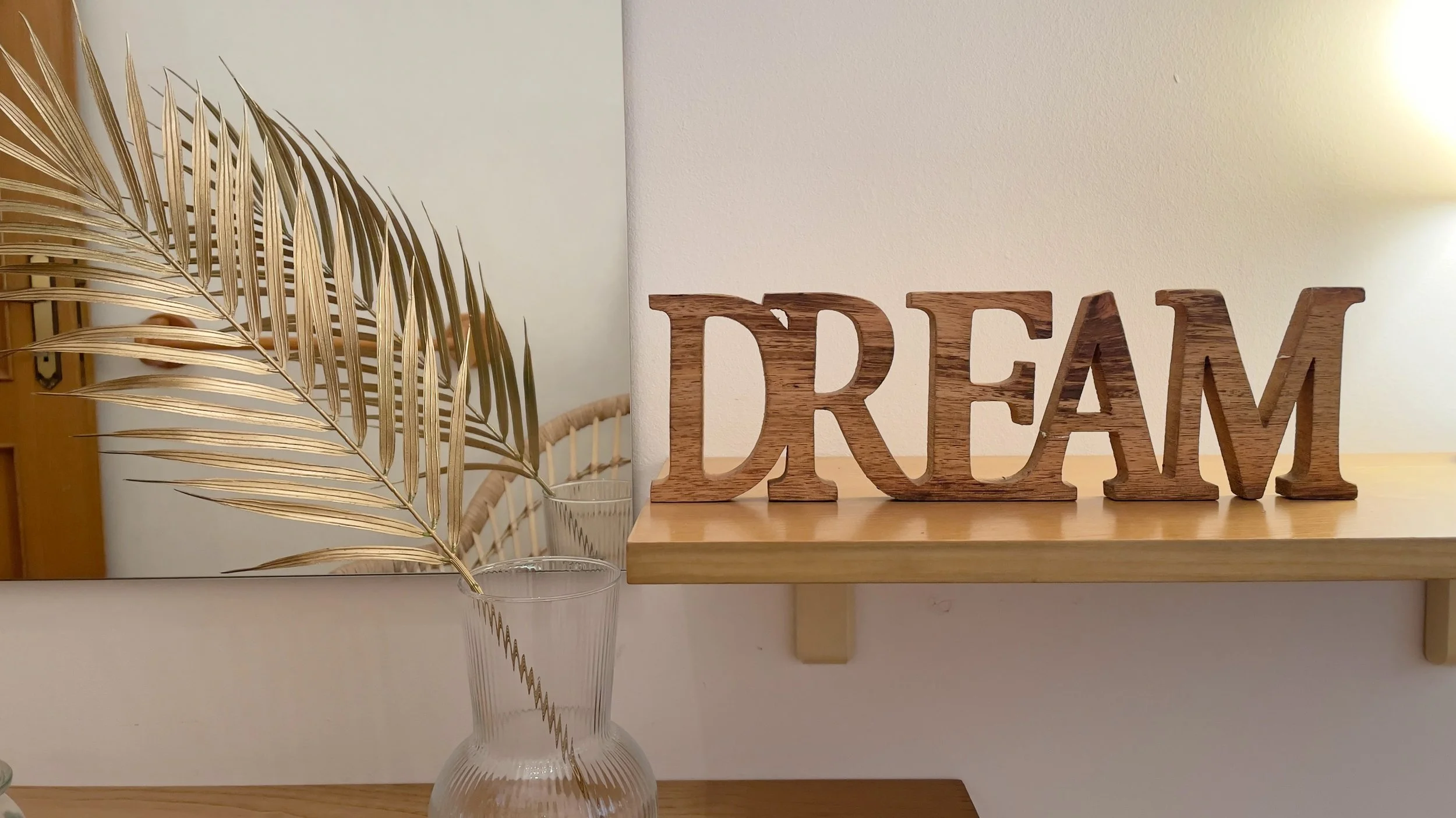 Decorazione da interni con foglia dorata in vaso di vetro trasparente e scritta "DREAM" in legno, posta su una mensola accanto a uno specchio.