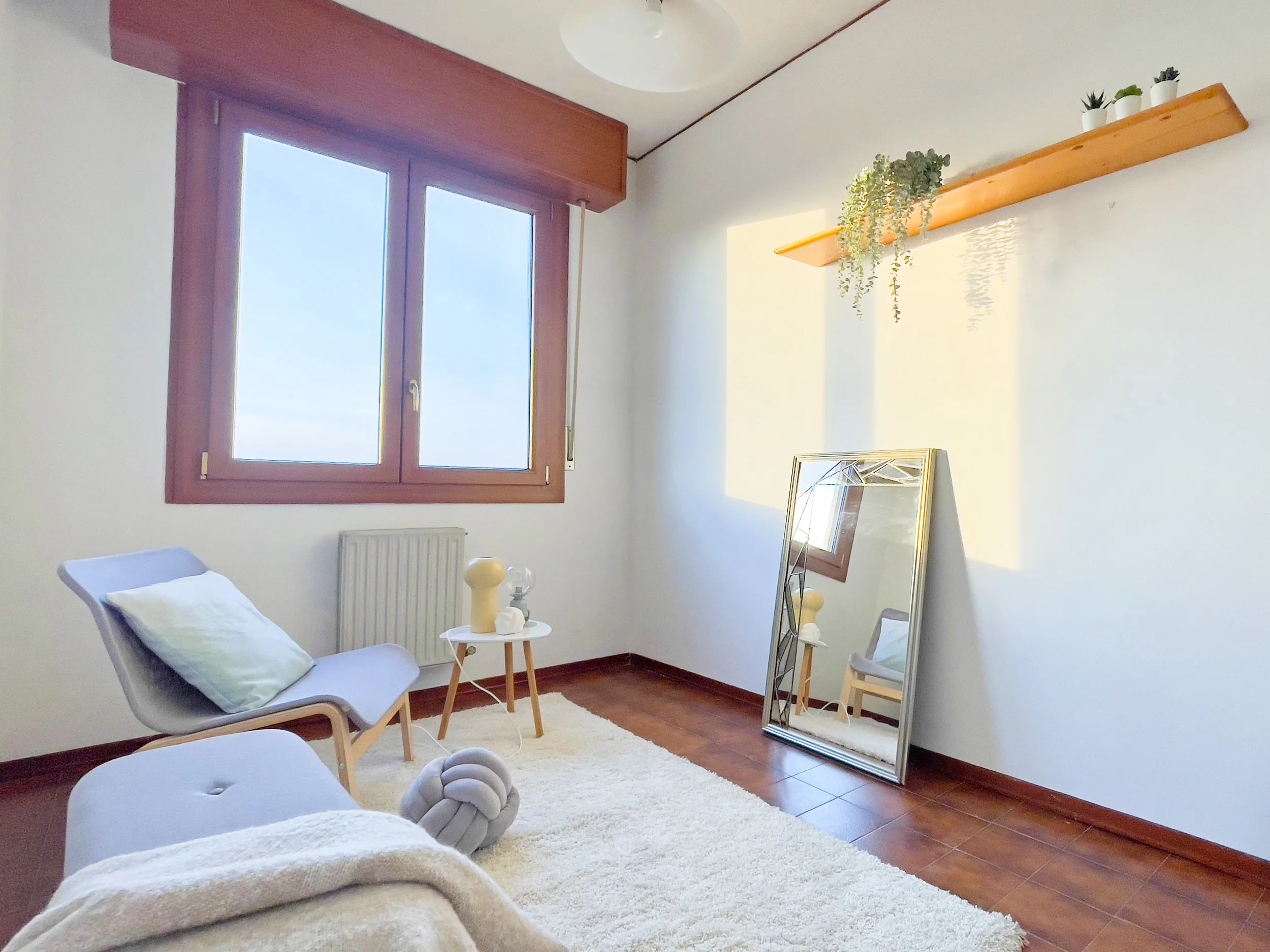 Stanza minimalista con una grande finestra, una sedia moderna, un tavolino con oggetti decorativi, uno specchio appoggiato al muro e una mensola con piante in vaso.