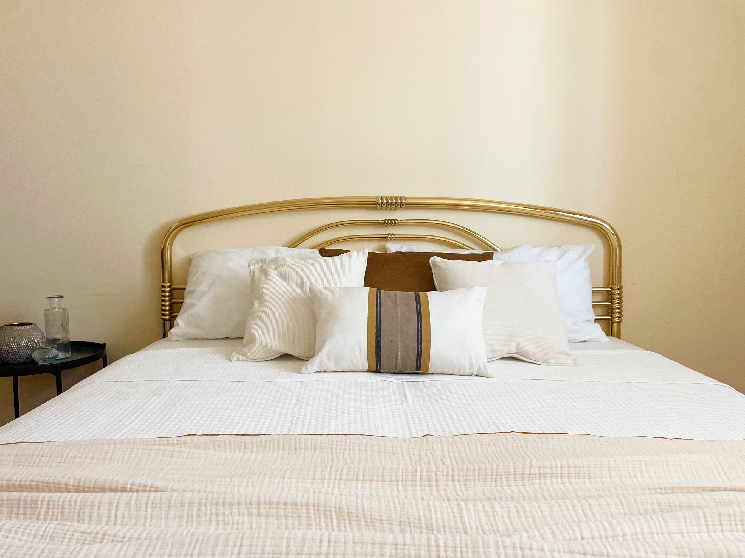 Letto con testiera dorata, cuscini bianchi e marroni, copriletto bianco, tavolino nero con decorazioni.