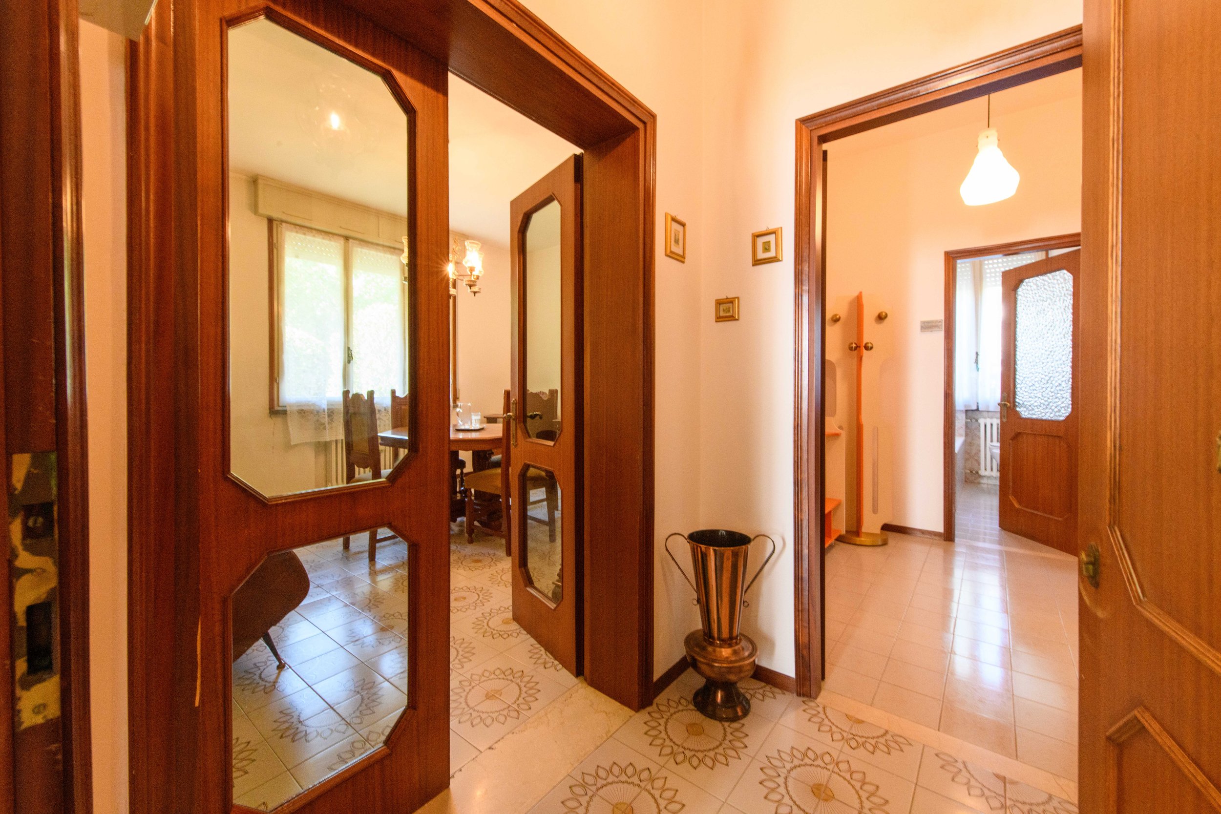 Interno di una casa con corridoio e porte in legno. A destra c'è un corridoio con una lampada a soffitto accesa. A sinistra si vede una sala da pranzo con un tavolo e sedie. Sul pavimento ci sono mattonelle con un motivo decorativo. Presente anche un