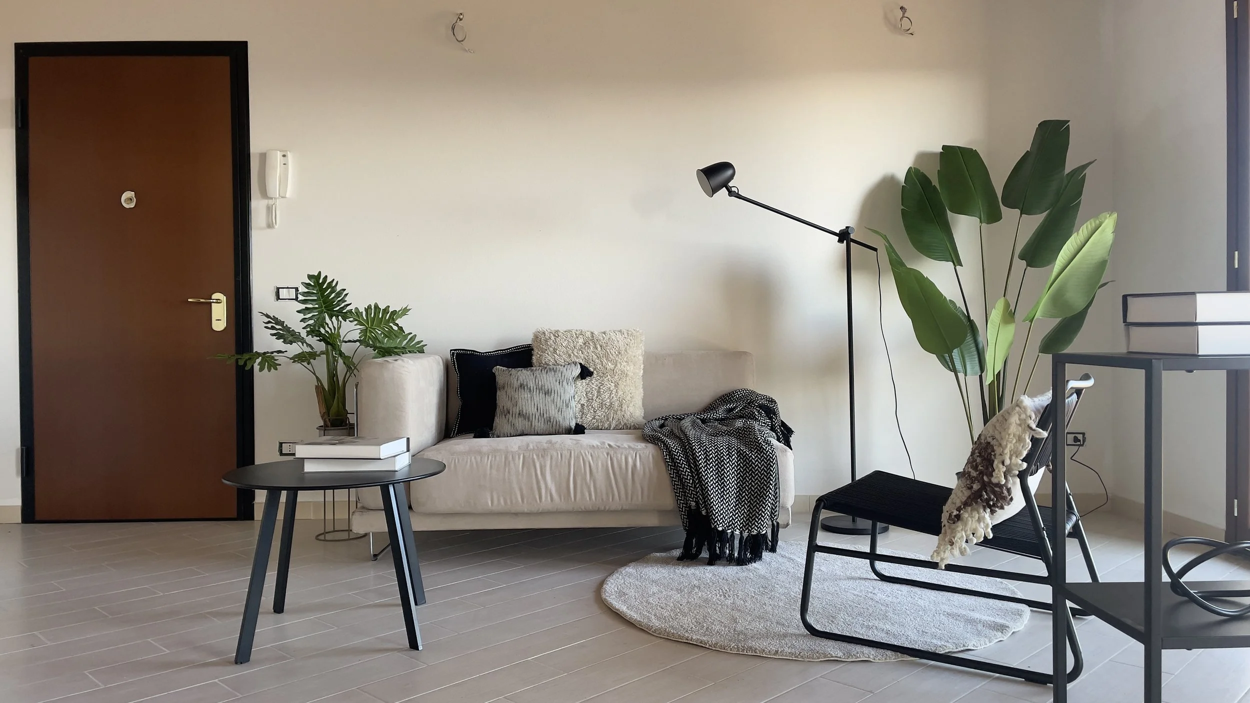 Soggiorno moderno con divano beige, cuscini, pianta tropicale, tavolino rotondo nero, lampada da terra, e sedia accanto a una porta di legno.