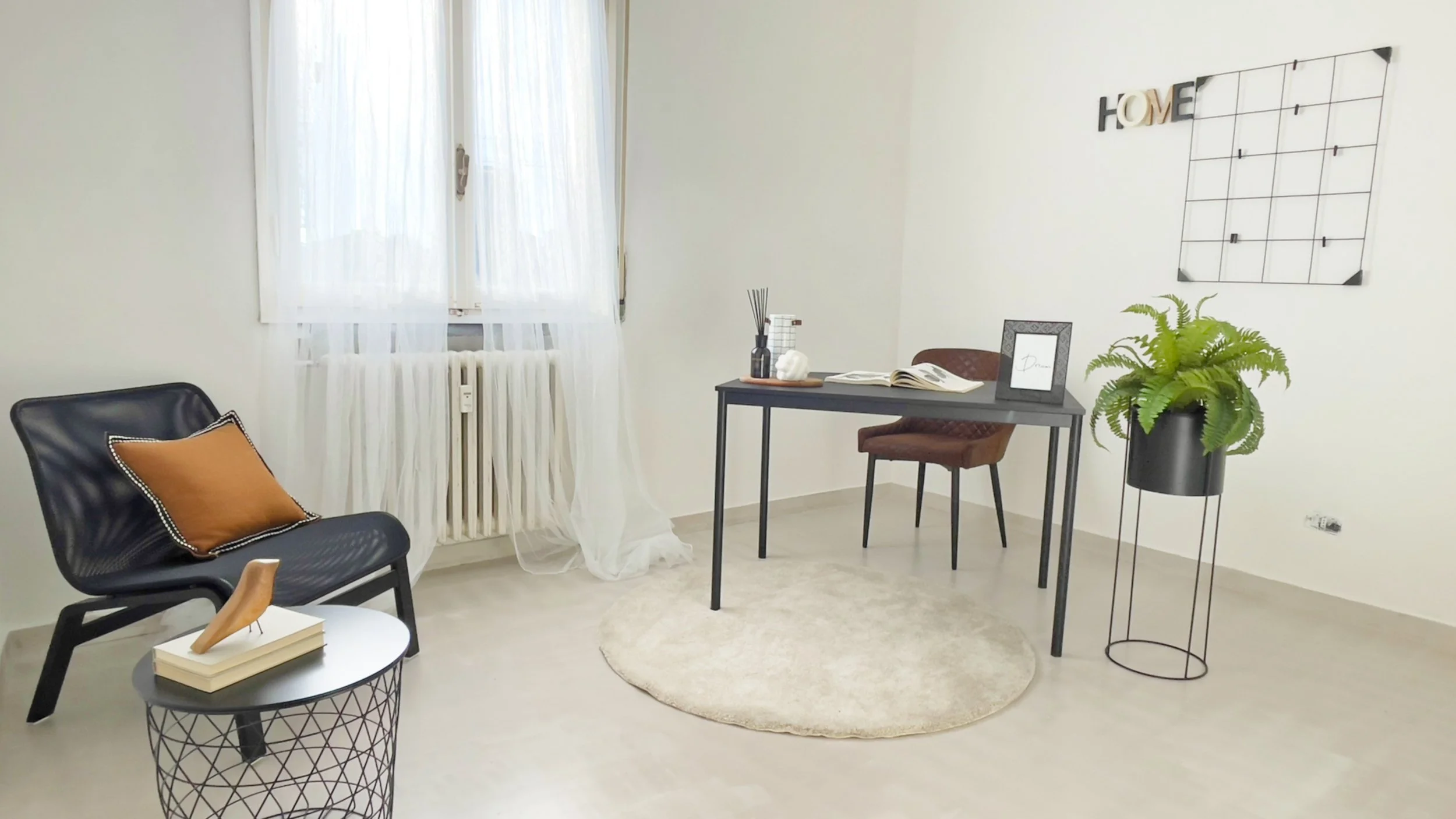 Interno di una stanza moderna con sedia nera e cuscino arancione, tavolino rotondo con oggetti decorativi, scrivania con accessori, pianta in vaso e finestra con tende bianche trasparenti.