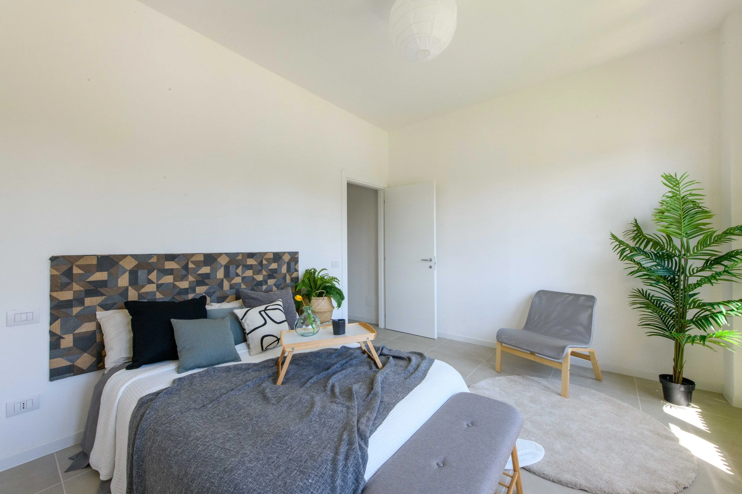 Camera da letto moderna con letto matrimoniale, cuscini decorativi, testiera geometrica in legno. Tavolino da colazione sul letto, pianta verde vicino a una poltrona grigia. Luce naturale, pareti bianche e arredamento minimalista.
