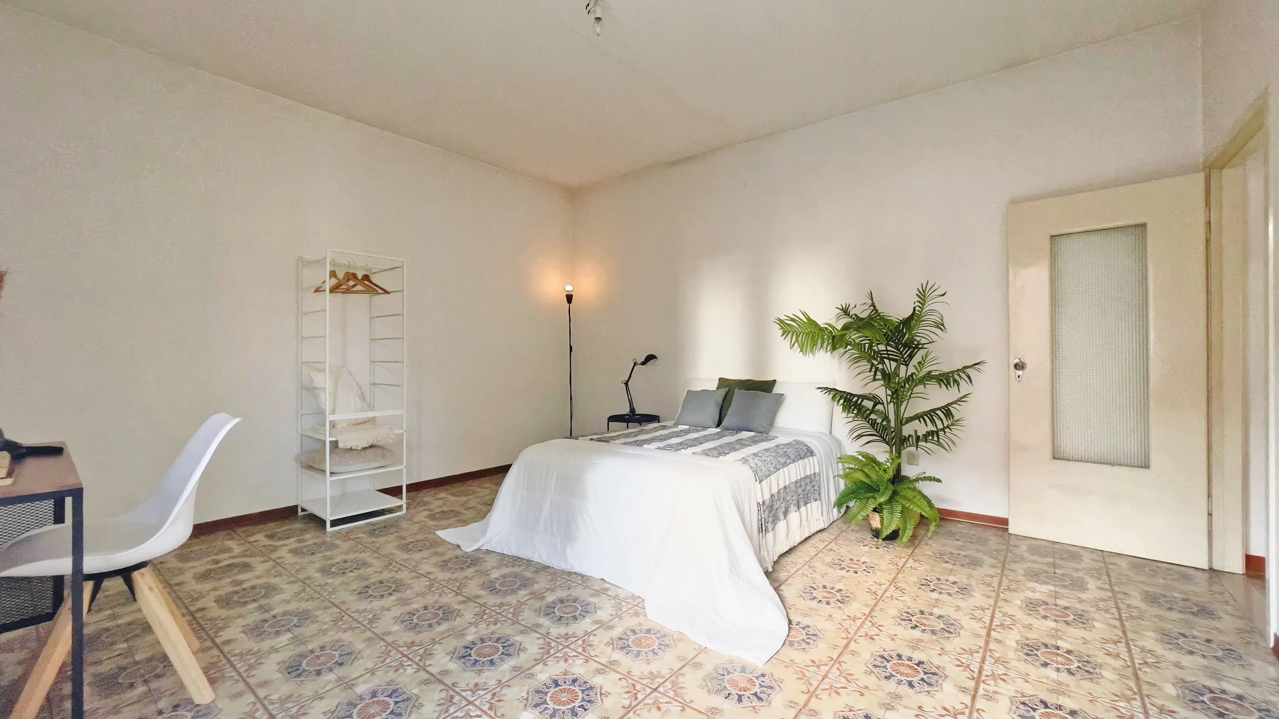 Camera da letto luminosa con letto matrimoniale, pianta verde accanto, lampada da terra, sedia bianca, scrivania nera e appendiabiti. Pavimento piastrellato decorativo.