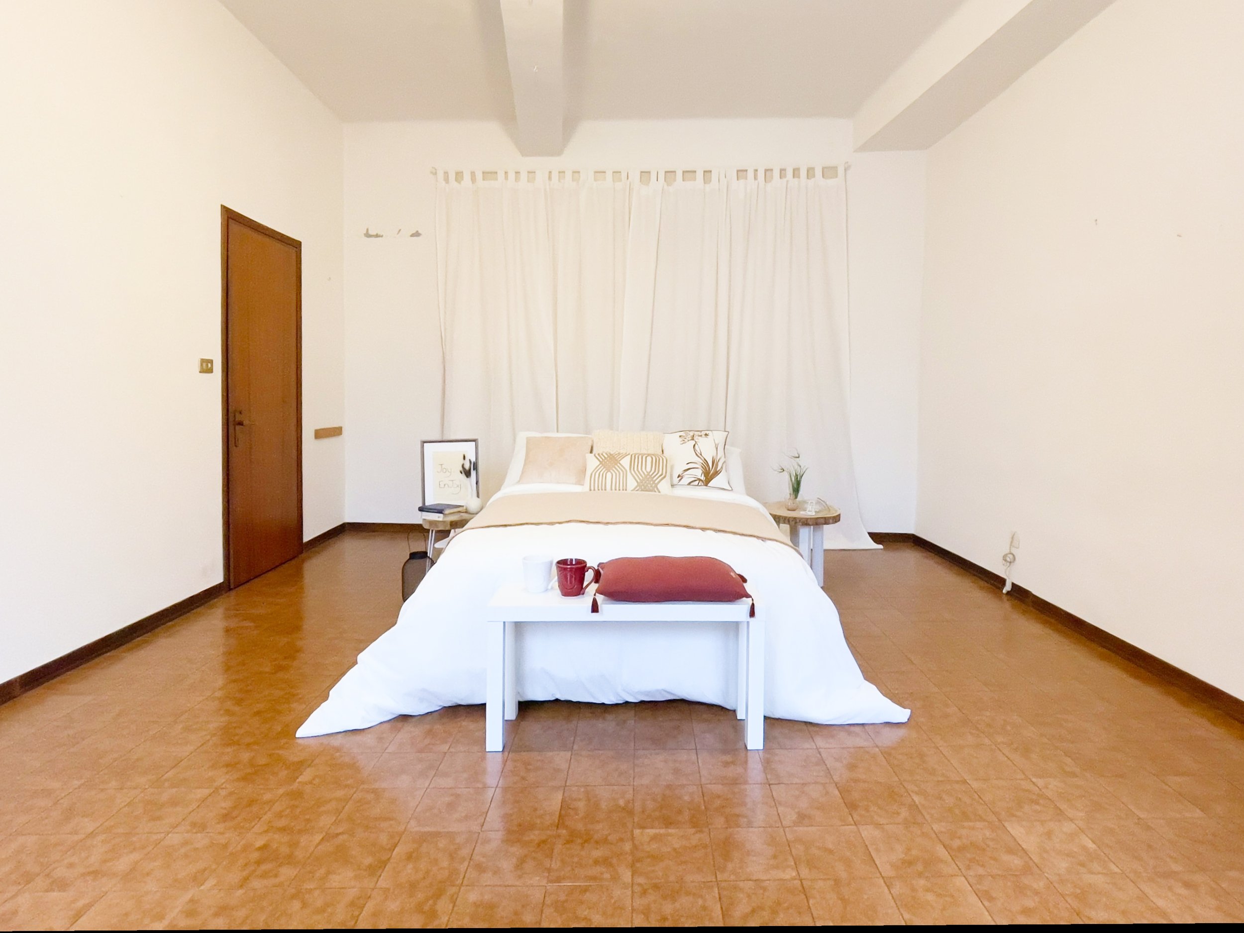 Camera da letto con letto matrimoniale, pavimento in piastrelle marroni, tende bianche, due comodini e tavolino con decorazioni.