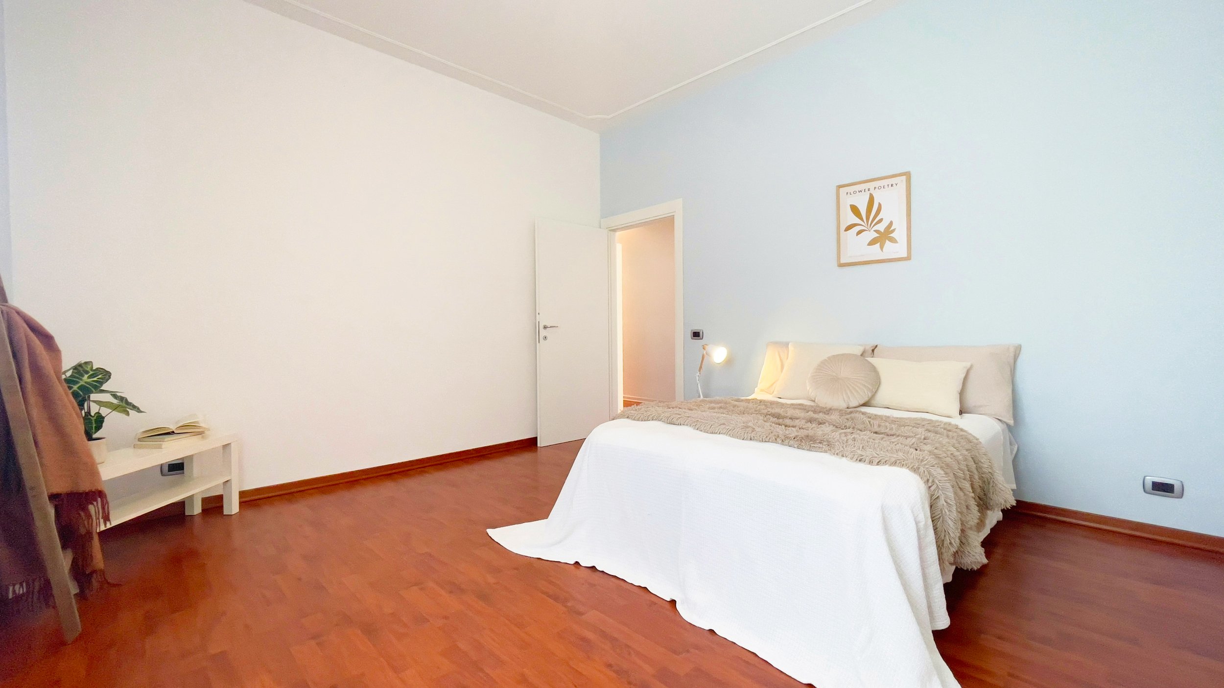 Camera da letto luminosa con letto matrimoniale, lenzuola bianche, cuscini decorativi e coperta marrone chiaro. A sinistra, una pianta su un tavolino vicino a una giacca appesa. Pareti chiare e pavimento in legno.
