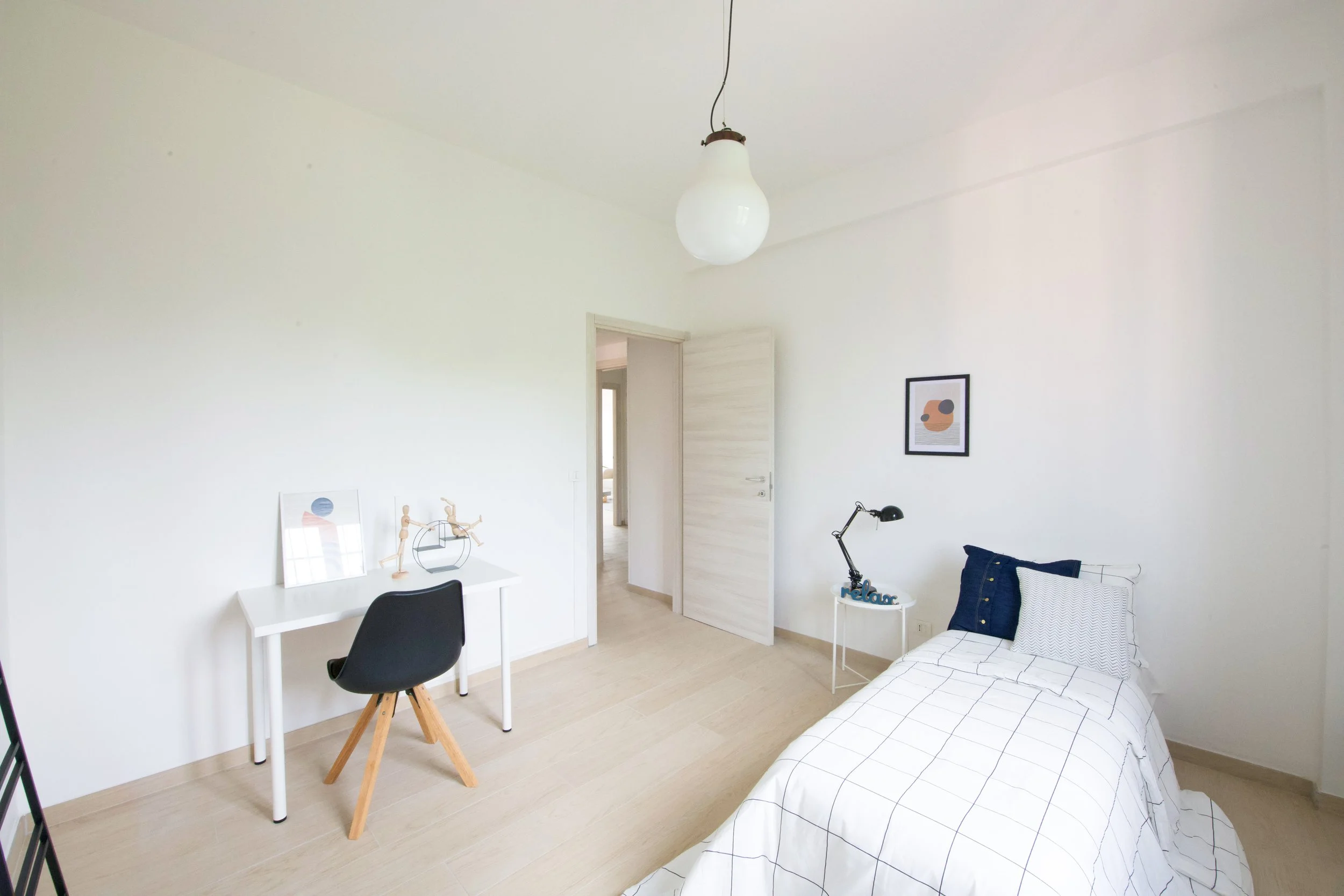 Camera da letto minimalista con pareti bianche, letto singolo con coperta a quadri, scrivania bianca e sedia nera, lampada da tavolo, decorazioni semplici, lampadario sospeso, e porta aperta.