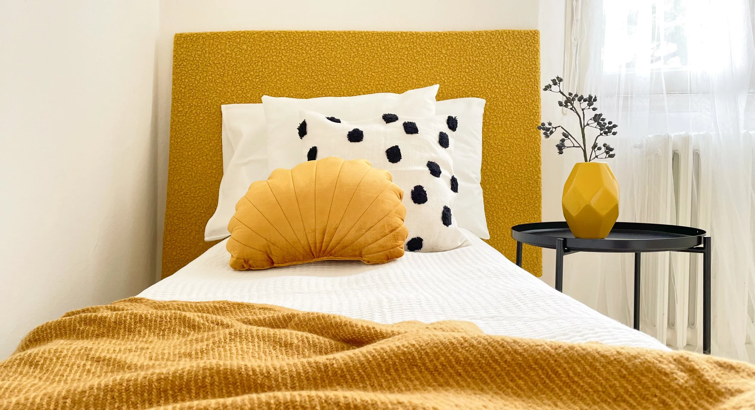 Camera da letto con letto singolo, coperta e cuscini gialli, cuscino bianco a pois neri, tavolino nero con vaso giallo e pianta decorativa; luce naturale entra dalla finestra.