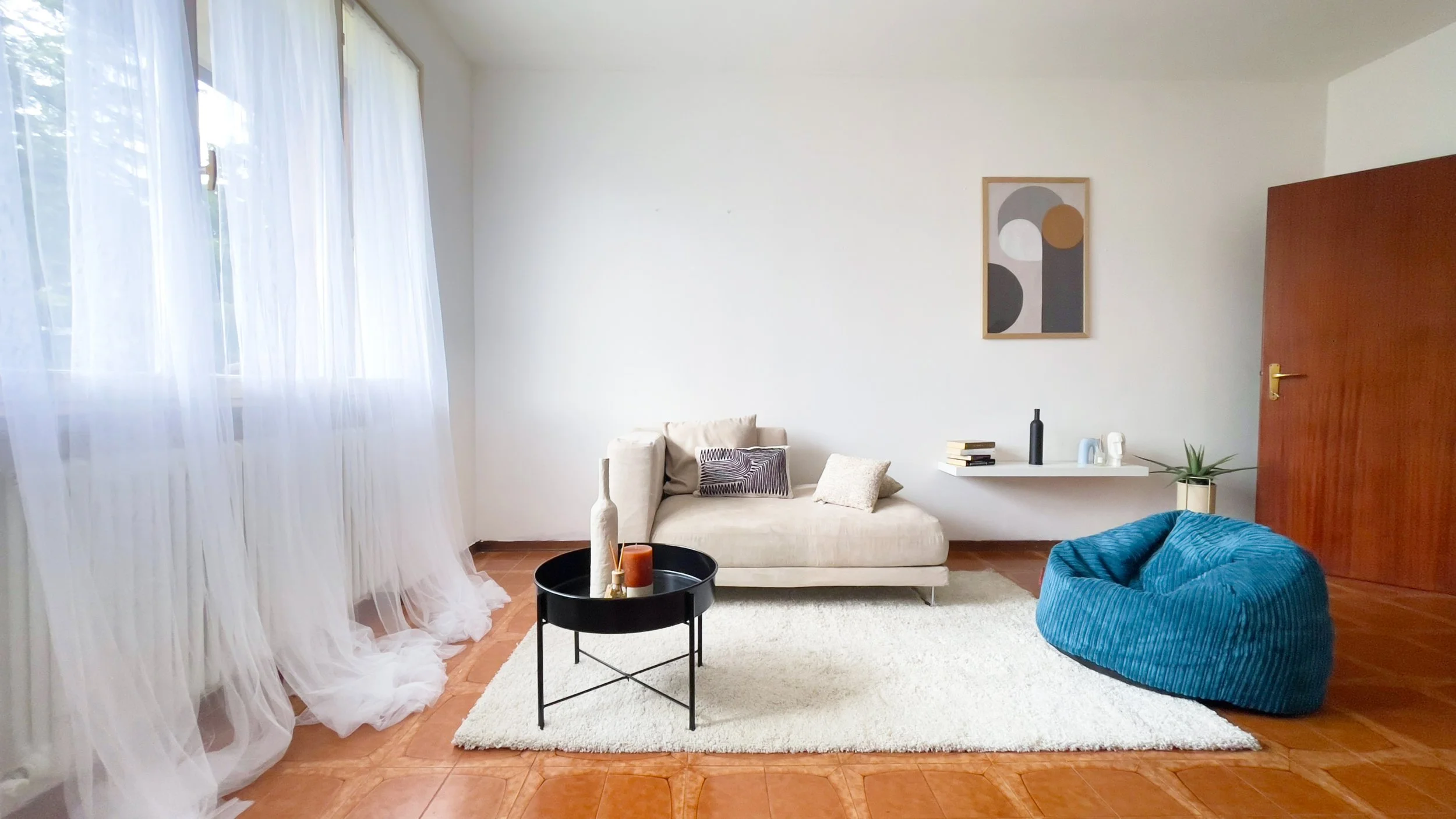 Soggiorno moderno con divano beige, tappeto bianco, tavolino nero e pouf blu. Parete con quadro astratto e mensola con libri e decorazioni. Luminosità naturale da grandi tende bianche.