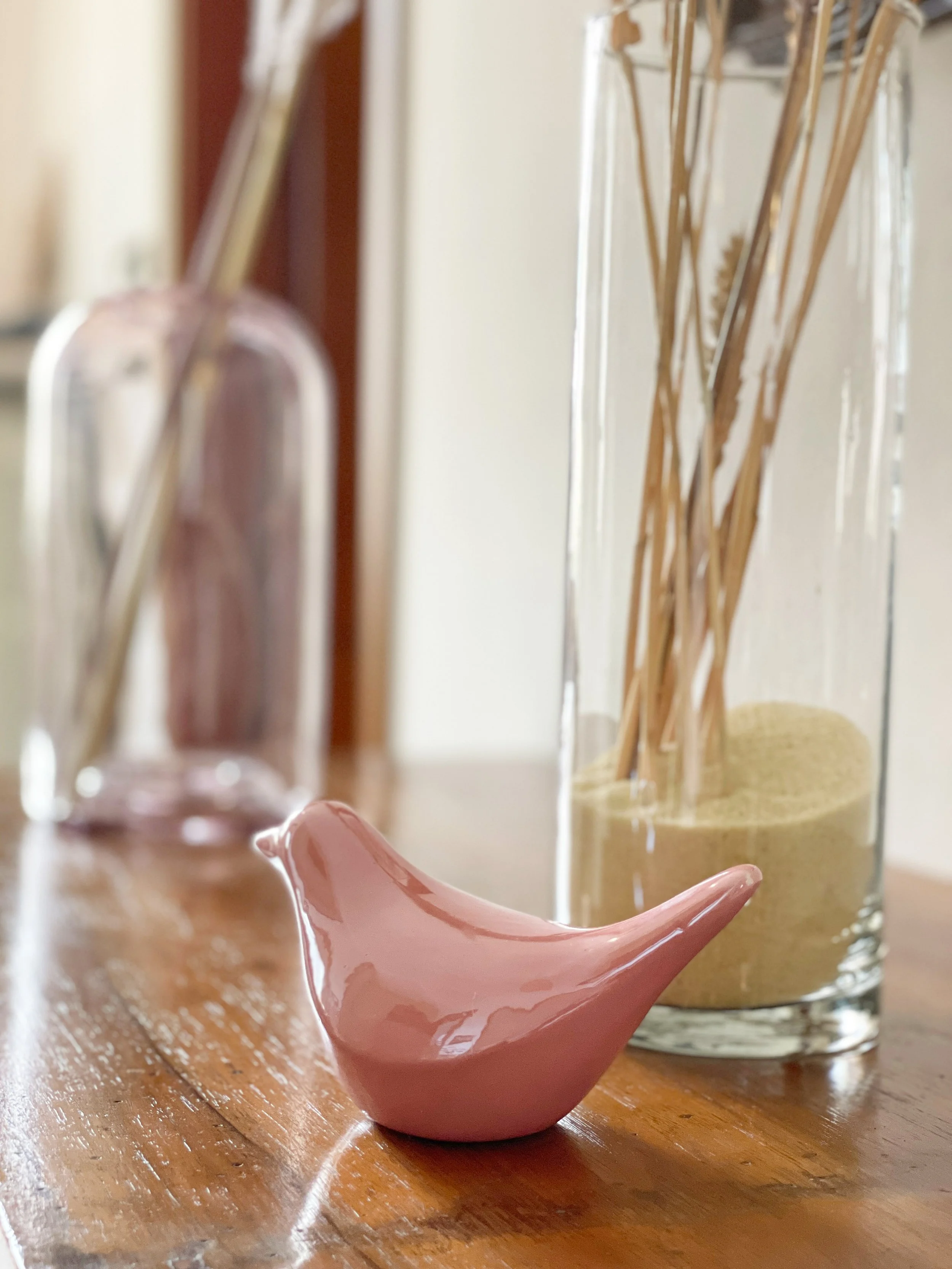 Decorazione d'interno con un uccellino di ceramica rosa su un tavolo di legno, accanto a due vasi di vetro con piante secche.