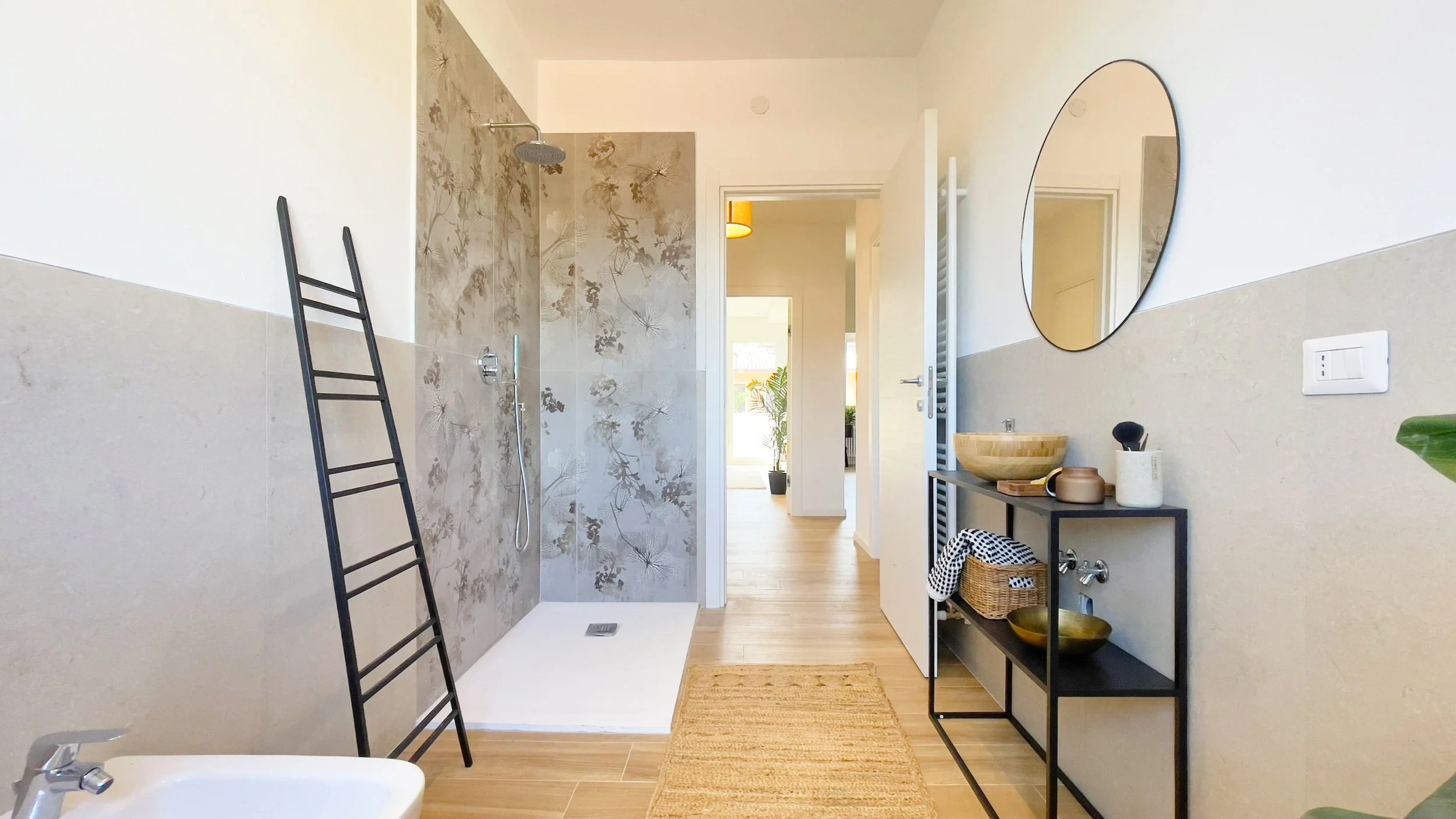 Bagno moderno con doccia aperta, pareti beige con decorazioni floreali, specchio rotondo, scaffale con accessori e pianta decorativa, porta aperta verso corridoio luminoso.