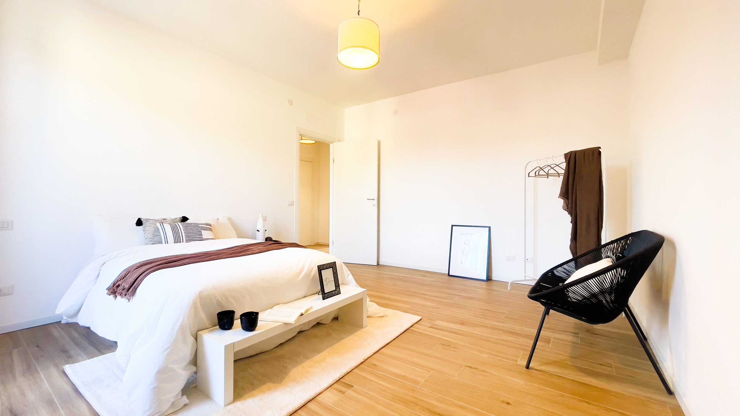 Camera da letto minimalista con letto matrimoniale, coperte bianche, cuscini decorativi, tavolino con tazze e libro, poltrona nera, appendiabiti con vestiti e pavimento in legno chiaro.
