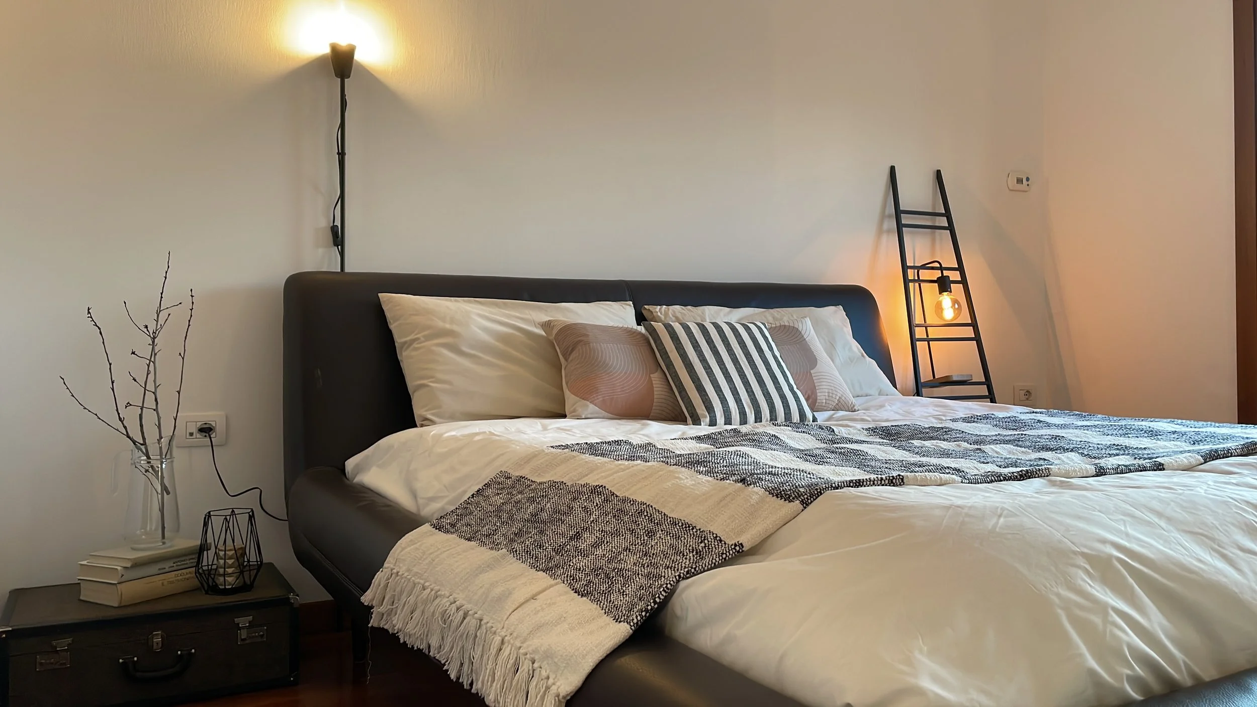 Camera da letto moderna con letto matrimoniale, cuscini decorativi, luce da parete, scala decorativa con lampada, e comodino con libri e rami secchi in vaso.