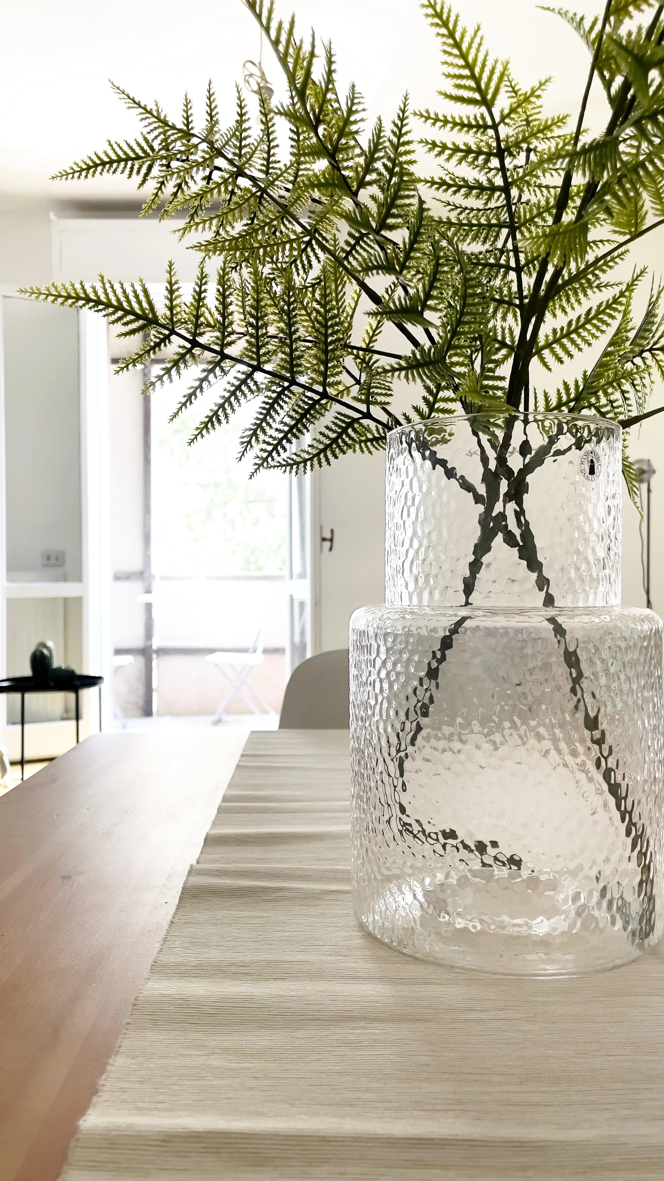 Vaso di vetro trasparente con foglie verdi su un tavolo di legno in un interno luminoso.