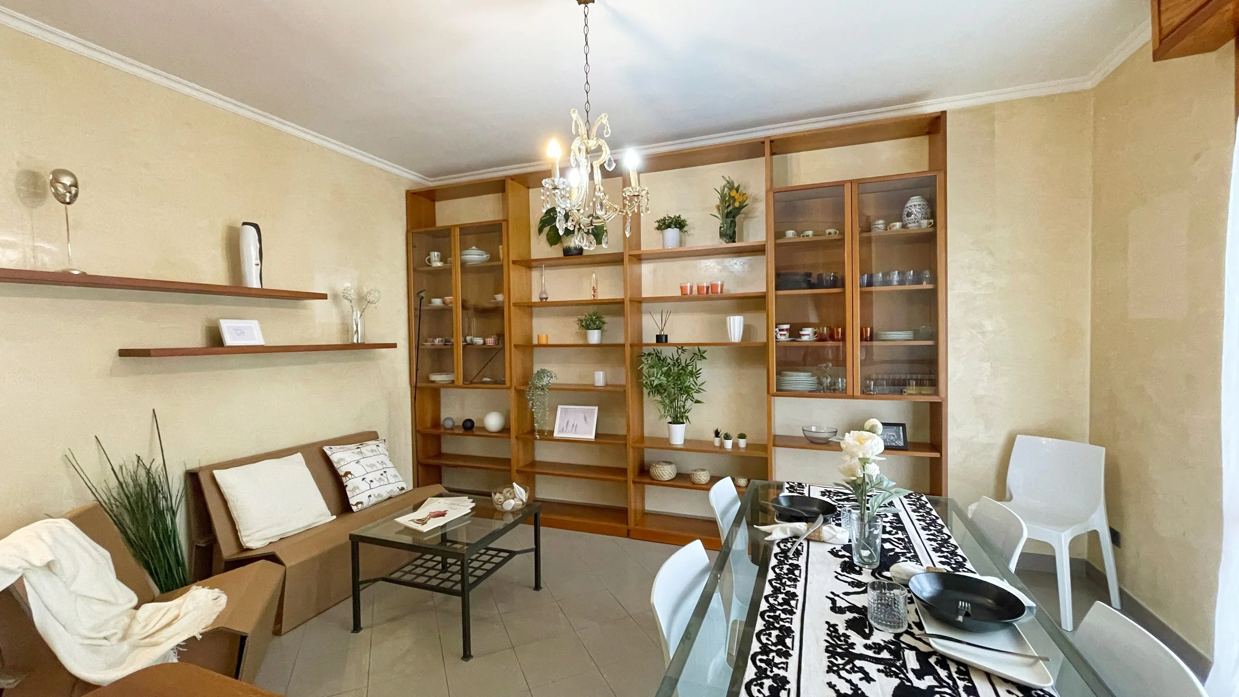 Soggiorno moderno con pareti beige, libreria a scaffali in legno, tavolo da pranzo con tovaglia bianca e nera, sedie bianche, divano con cuscini bianchi e neri, piante decorative e lampadario sopra il tavolo.
