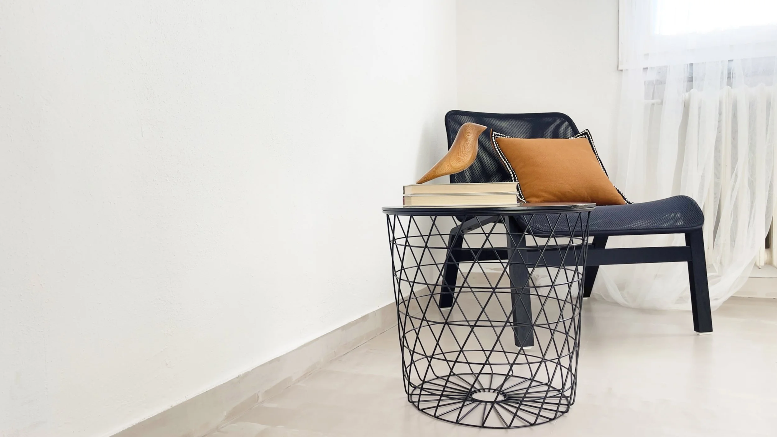 Sedia nera con cuscino arancione, tavolino in metallo con libri e decorazione in legno, stanza bianca con tenda.