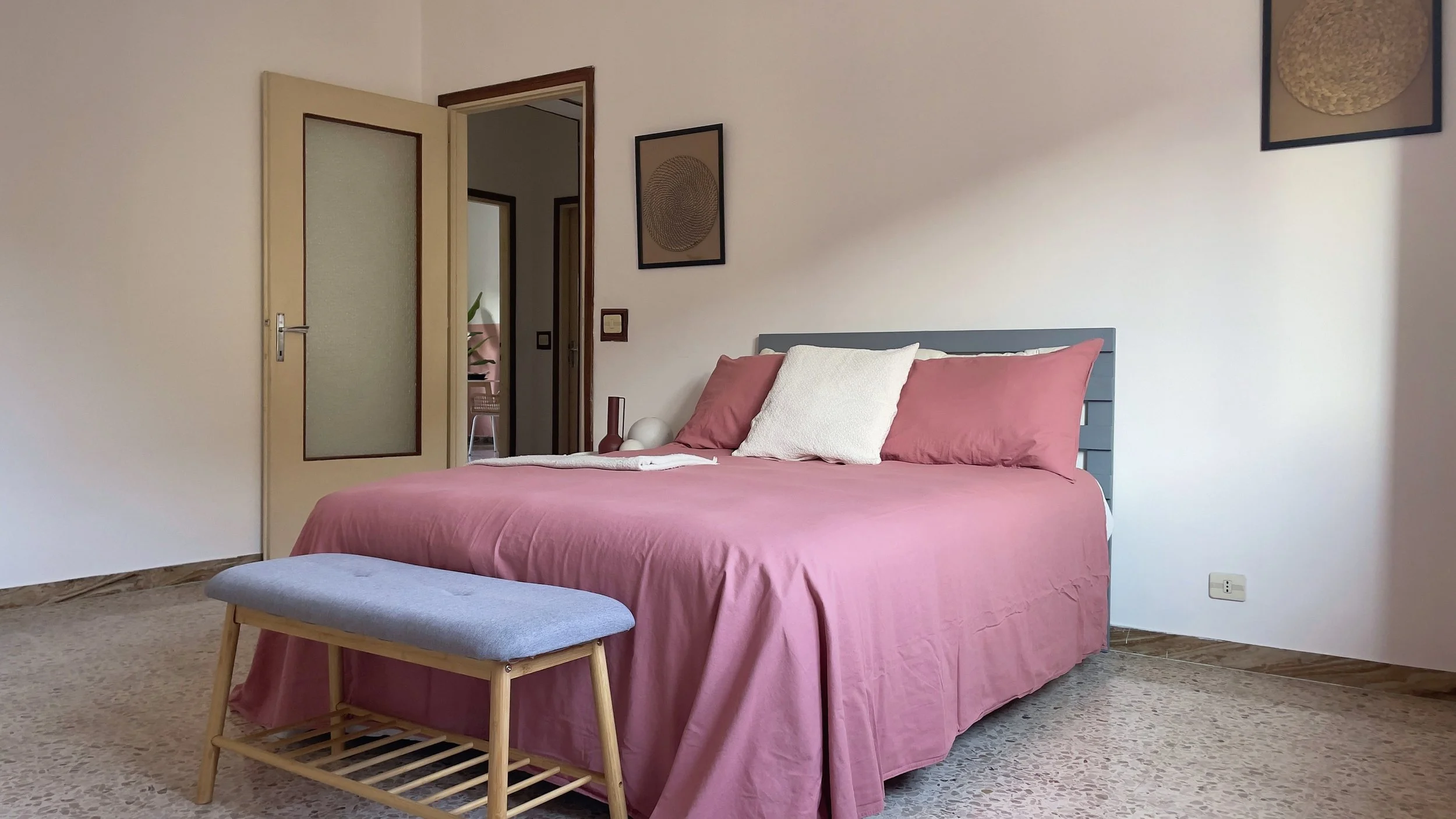 Camera da letto con letto matrimoniale rosa, panca ai piedi, pavimento in marmo, porta aperta e due quadri appesi.