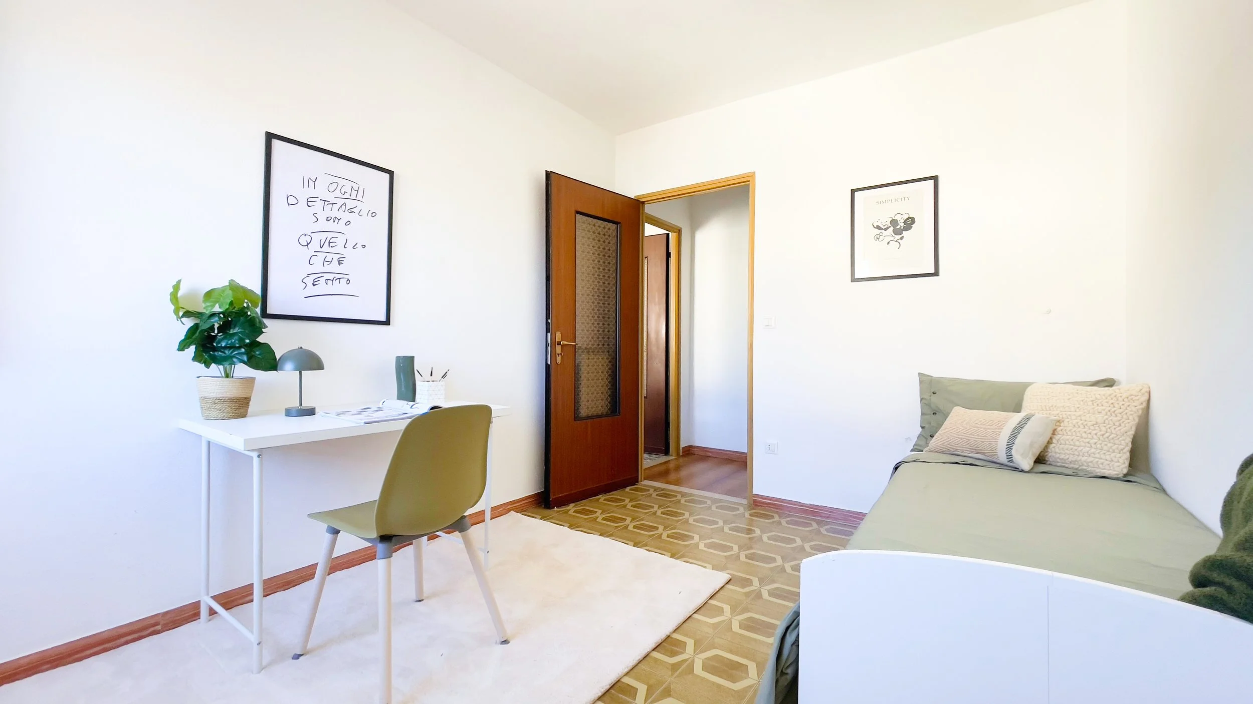 Cameretta moderna con una scrivania bianca, sedia verde, pianta in vaso, lampada da tavolo, letto singolo con cuscini, e porta in legno aperta. Pareti bianche decorate con quadri.
