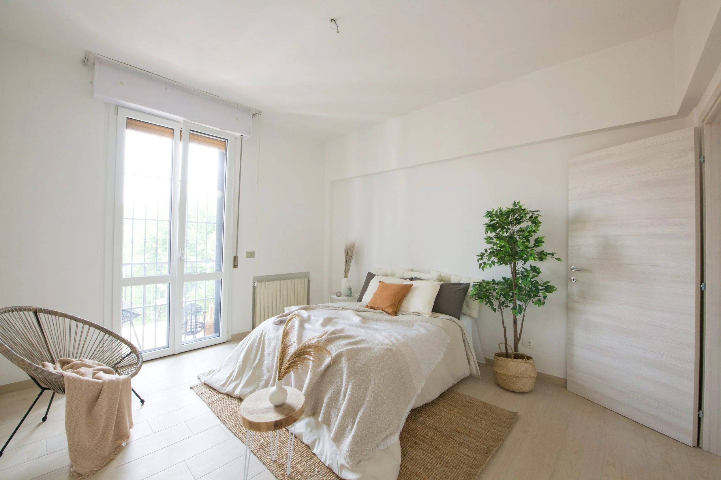 Camera da letto moderna con letto matrimoniale, pareti bianche, finestre alte con tendine, sedia rotonda con coperta, pianta decorativa in vaso, tappeto in fibra naturale e tavolino di legno.