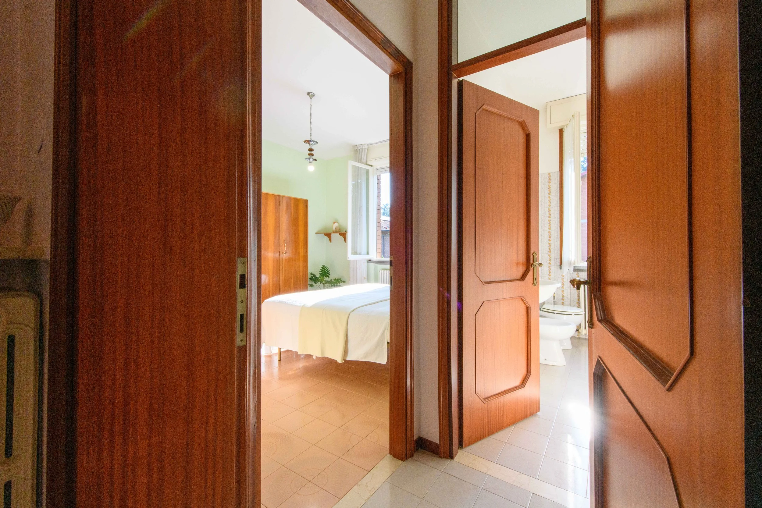 Vista di un corridoio con porte aperte che conducono a una camera da letto luminosa e un bagno. La camera ha pareti verdi, un armadio di legno e una finestra aperta. Il bagno è parzialmente visibile con un lavabo e una finestra sulla destra.