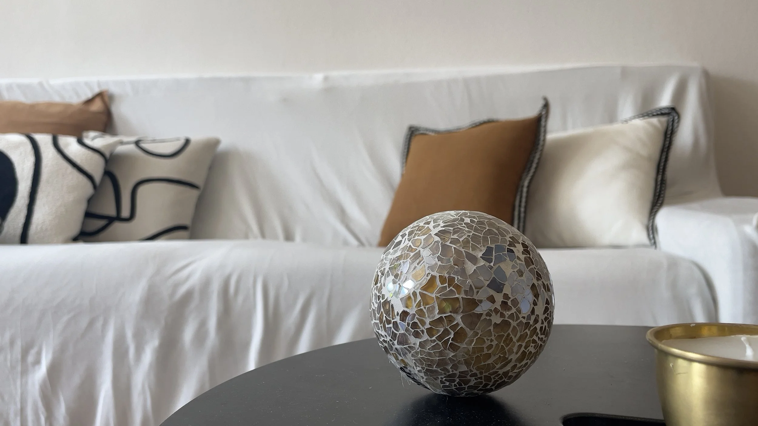 Interno di un salotto con divano bianco e cuscini decorativi, sfera ornamentale con mosaico su tavolino, tonalità beige e oro, ambiente accogliente.