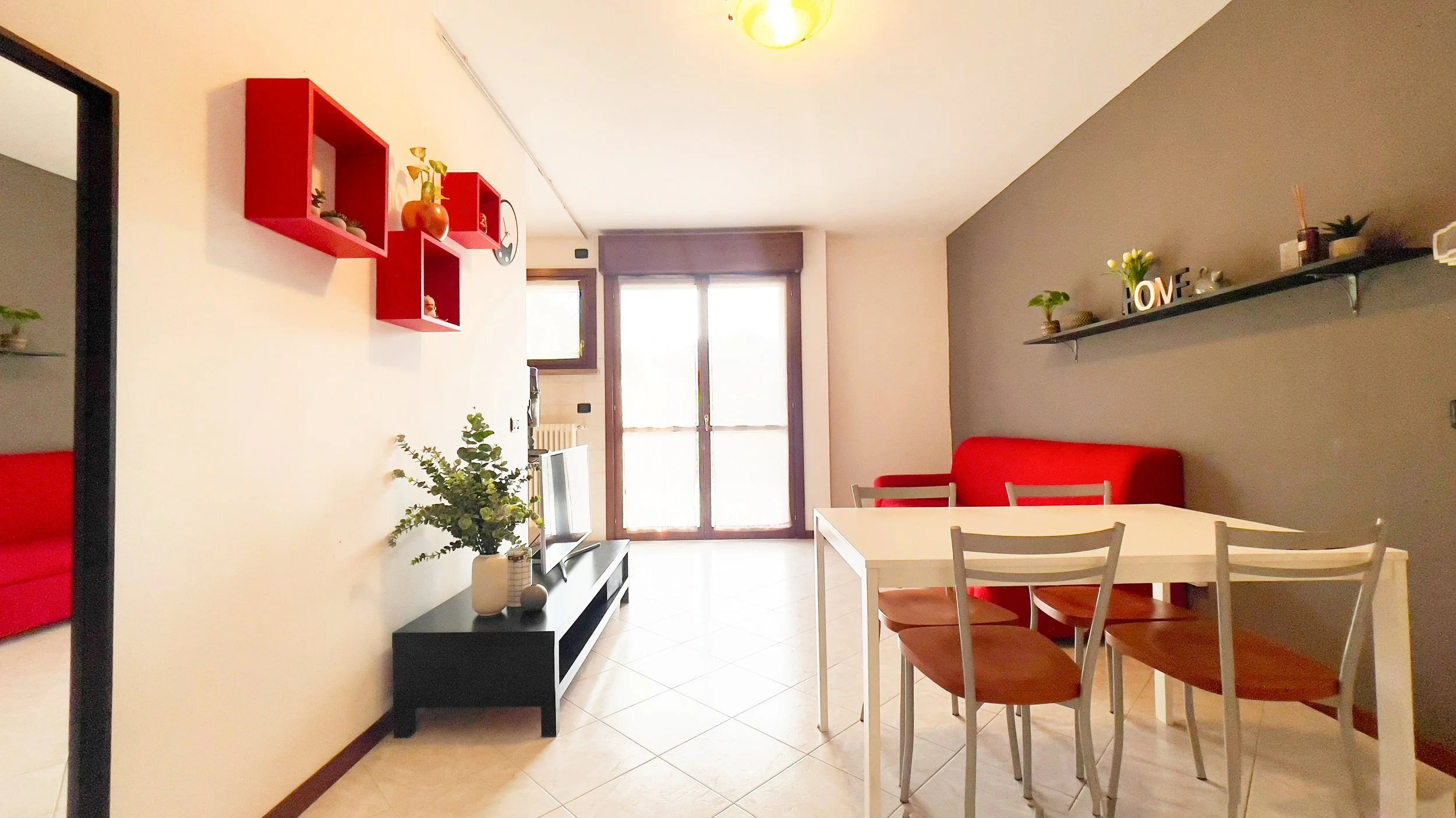Soggiorno moderno con tavolo da pranzo bianco e sedie, divano rosso, mensola con decorazioni, e finestre luminose.