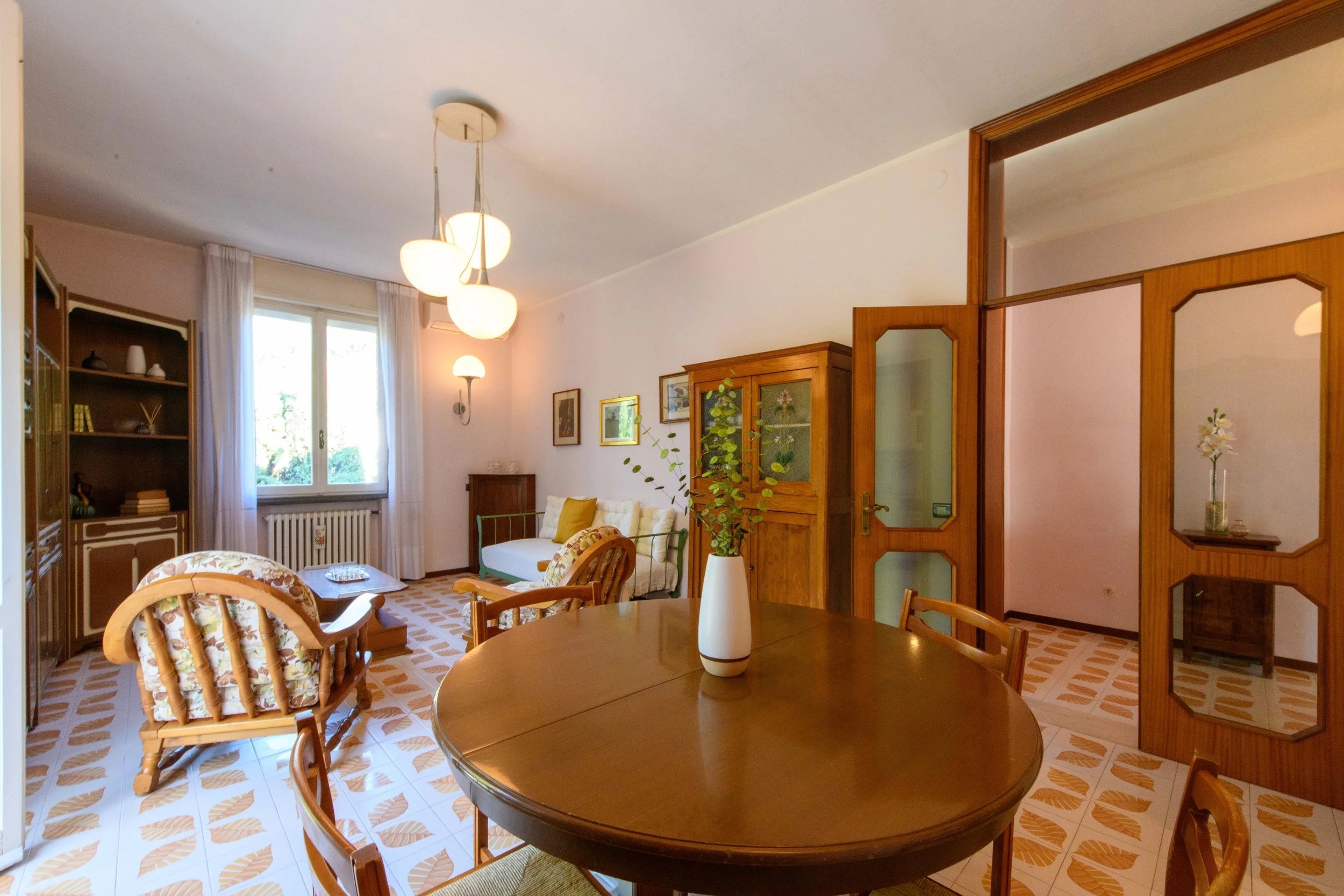 Salotto con arredamento vintage, tavolo rotondo in legno, soprammobili classici e lampadario a sospensione. Vista di una finestra con tende, poltrone e divano.