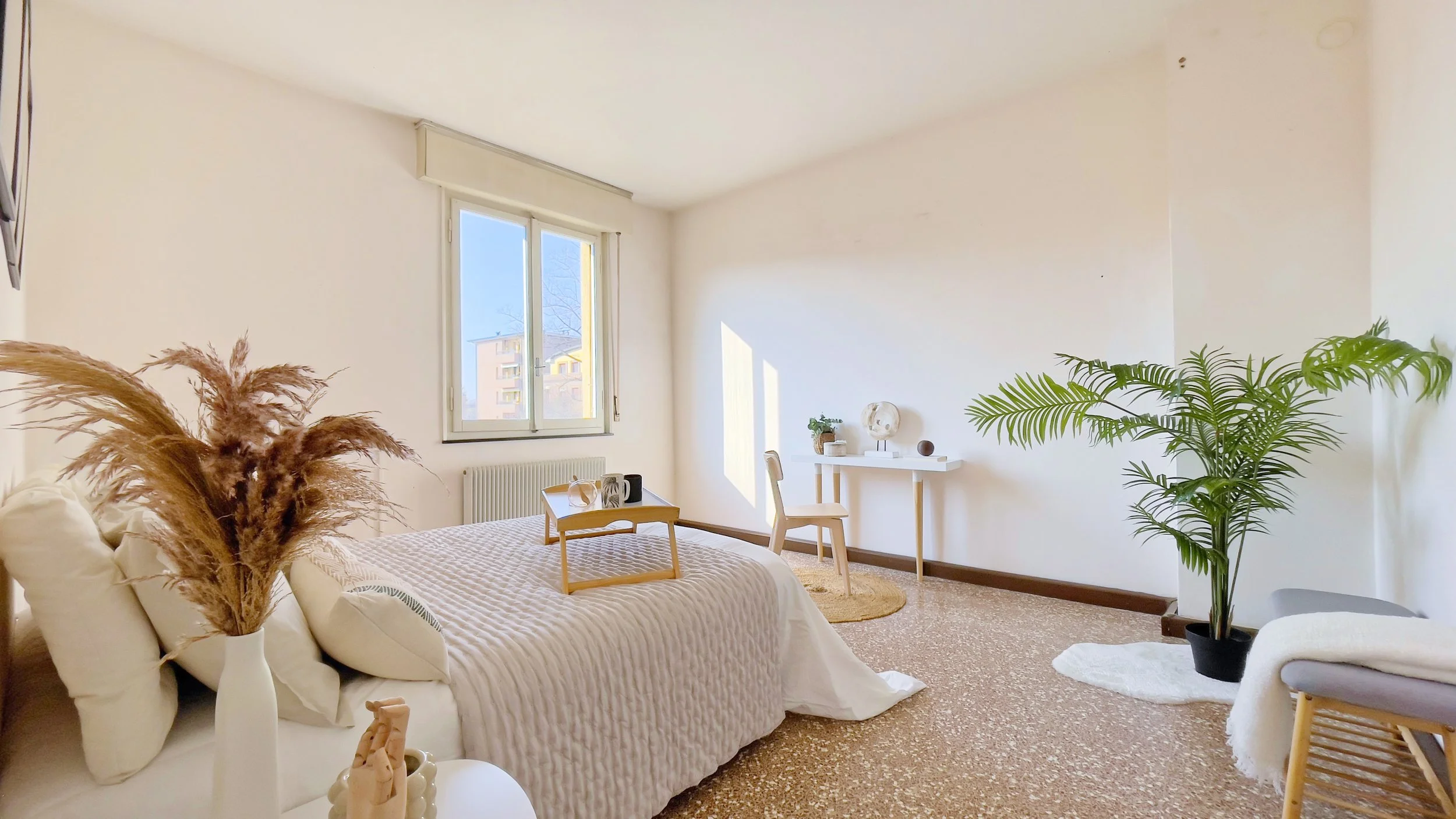 Camera da letto luminosa con letto matrimoniale, decorazioni minimaliste, tavolino, pianta verde e scrivania vicino alla finestra.