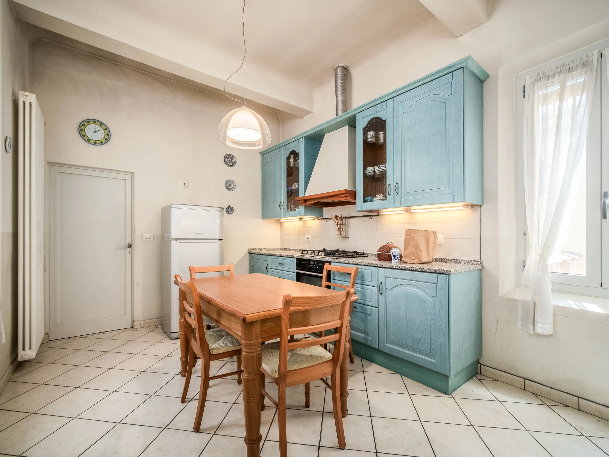 Cucina accogliente con mobili in legno chiaro, elettrodomestici moderni e pavimento in piastrelle bianche. Tavolo e sedie in legno al centro, mensole verdi con stoviglie, e una finestra che illumina l'ambiente.