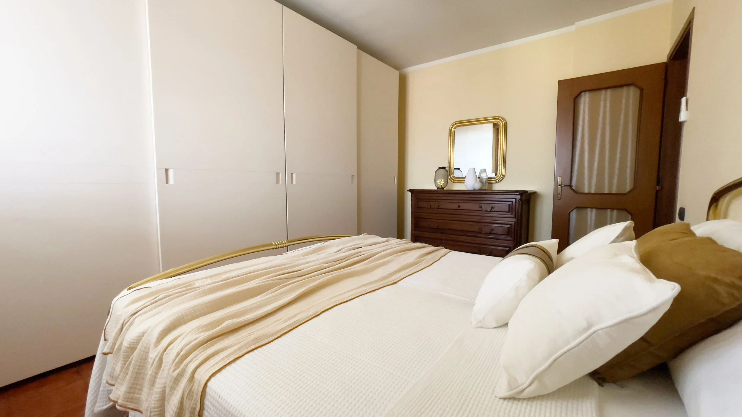 Camera da letto con letto matrimoniale, cuscini bianchi e marroni, grande armadio bianco, cassettiera in legno con specchio dorato, porta in legno.