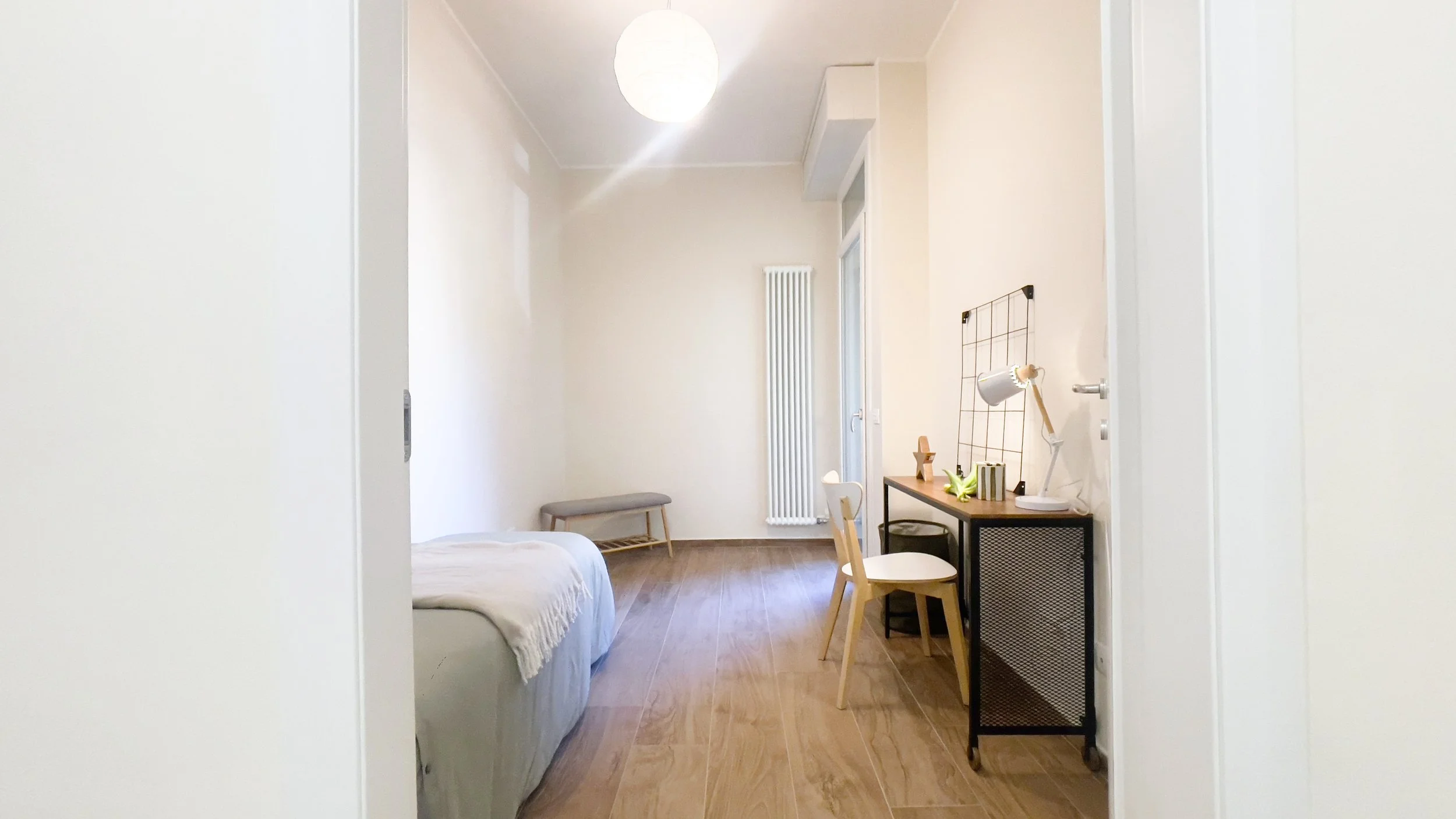 Interno di una camera da letto semplice e moderna, con pavimento in legno, un letto singolo con coperta beige, e un piccolo scrittoio con sedia e lampada da tavolo. Sullo sfondo è visibile un radiatore e una panca.