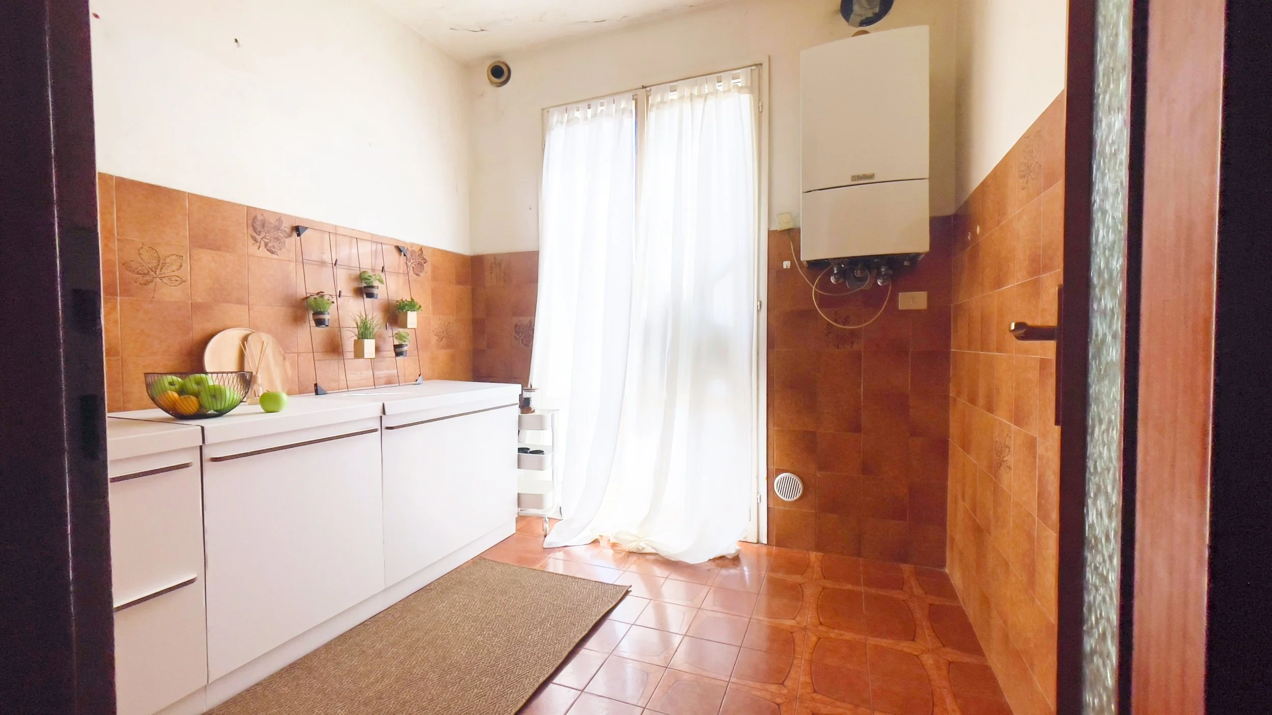 Cucina con pareti piastrellate marroni, un mobile bianco con ciotola di frutta, e piante appese. Una caldaia a muro e una porta finestra con tende bianche.
