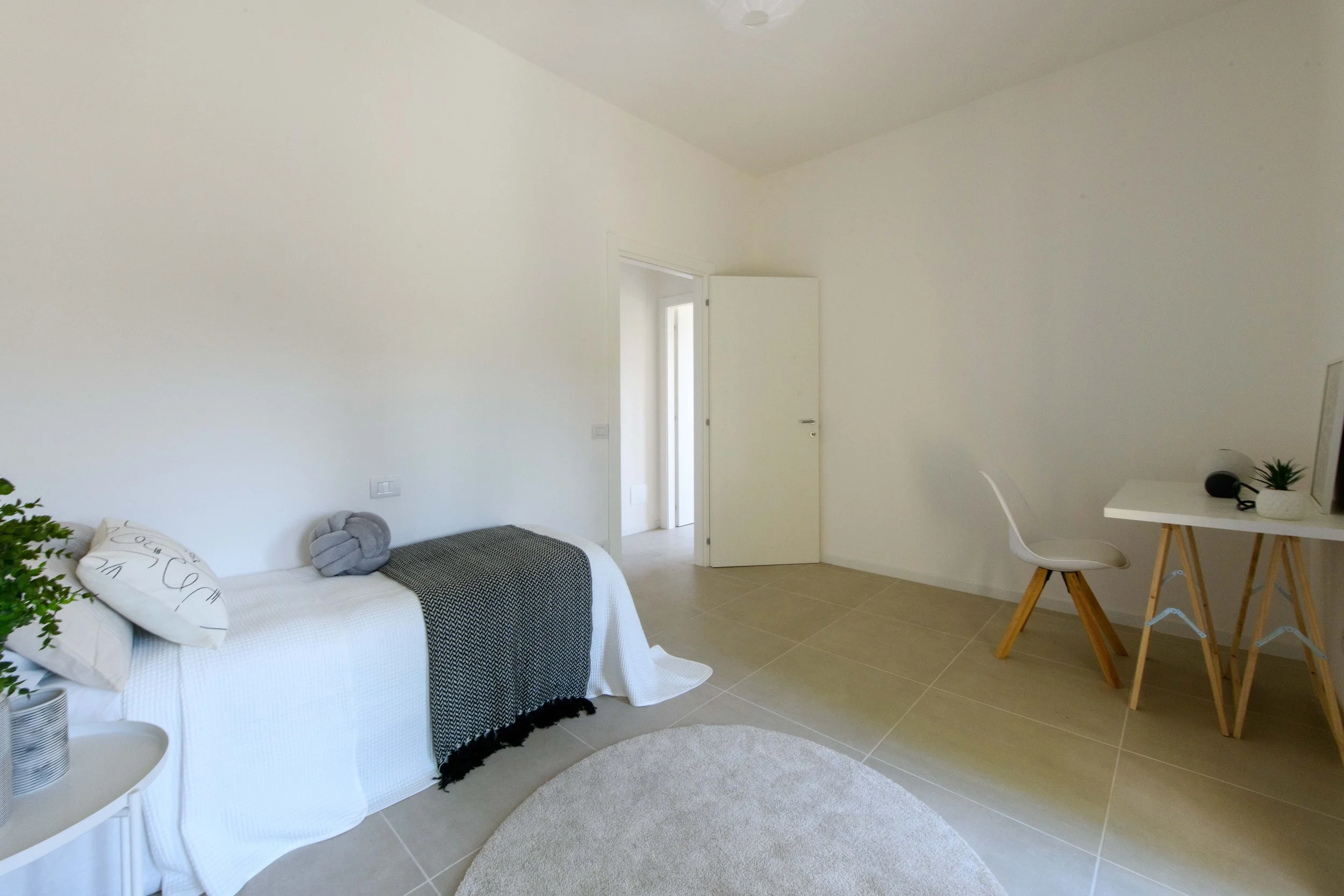 Camera da letto minimalista con letto singolo, coperta bianca, cuscini, tavolo bianco con pianta in vaso e sedia bianca, porta aperta, pavimento in piastrelle beige e pareti bianche.