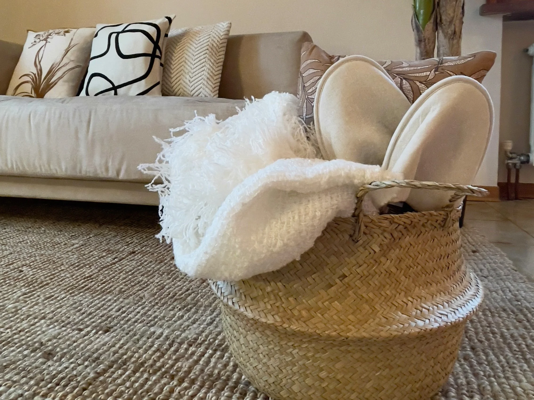 Interno di un salotto con un divano beige decorato con cuscini e un cesto di vimini contenente un plaid bianco e pantofole su un tappeto di juta.