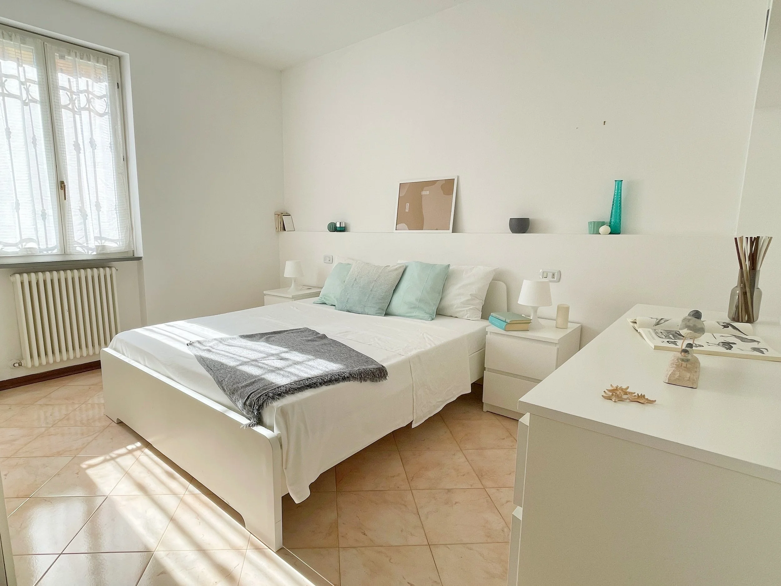 Camera da letto luminosa con letto matrimoniale, cuscini azzurri, coperta grigia e comodini bianchi. Ampia finestra con tende leggere, pavimento in piastrelle beige e decorazioni minimaliste su mensola e cassettiera.