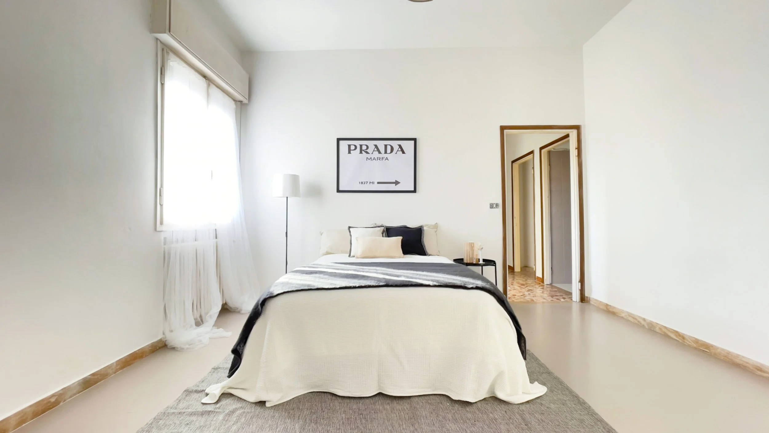Camera da letto minimalista con letto bianco, coperta grigia, lampada da terra e quadro "Prada Marfa" sulla parete, finestra con tende trasparenti, e pavimento beige.